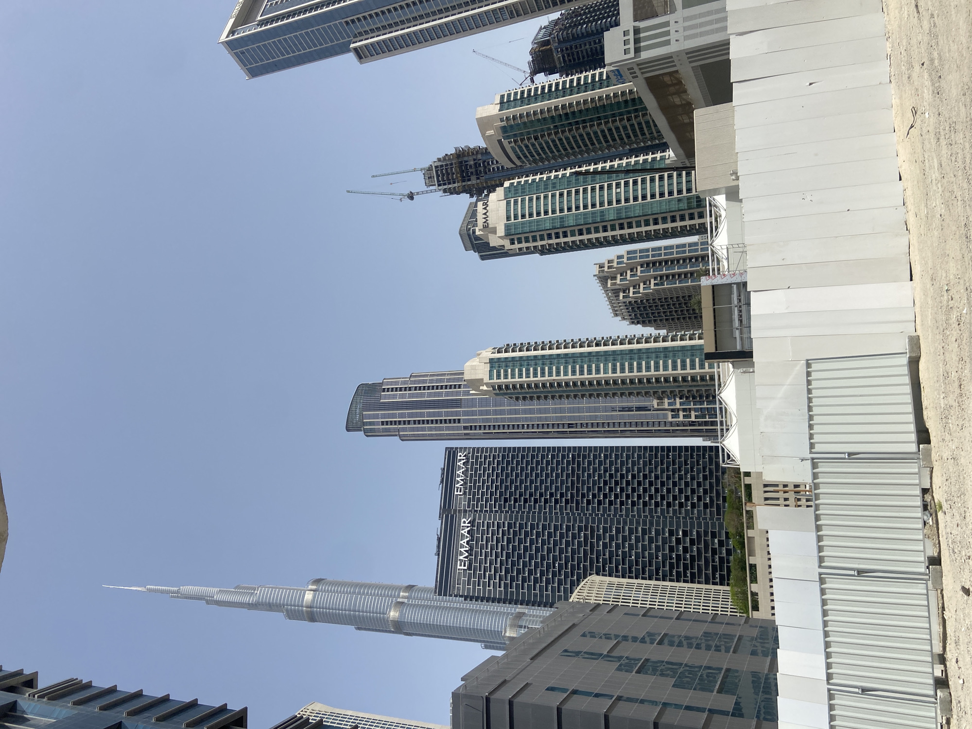 The image shows a cluster of skyscrapers in Dubai, United Arab Emirates, under a clear blue sky. The Burj Khalifa, the world's tallest building, is prominently visible, piercing the sky with its distinctive spire. Adjacent to the Burj Khalifa is a dark-colored skyscraper displaying the "EMAAR" logo. Other skyscrapers of varying designs and heights are arranged in the background, some with visible construction cranes. A row of white, corrugated metal structures runs parallel to the skyscrapers, suggesting a construction site or temporary barriers. The ground surface next to these metal structures appears to be a mix of light-colored dirt and concrete. No people are visible in the image.