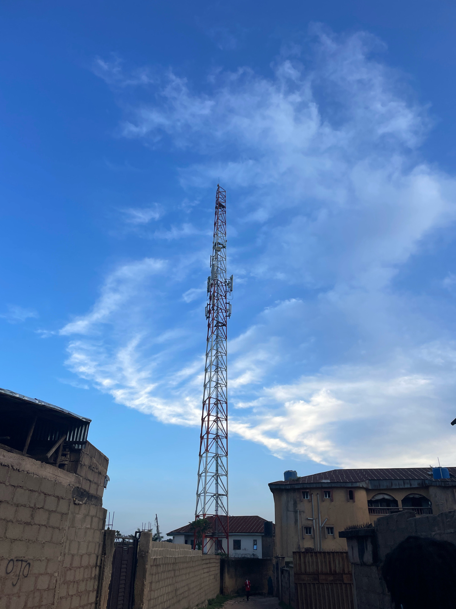 In Jos, Nigeria, a towering telecommunications tower dominates the sky, its lattice structure painted white with red accents. The tower is positioned amidst urban structures, with a weathered brick wall and a corrugated metal roof visible on the left, and a multi-story building with a sloped, metallic roof on the right. The sky is a vibrant blue, dotted with wispy white clouds. A narrow dirt path runs between the buildings, and a solitary figure in a red garment walks away from the camera in the distance. Several metal gates and fences are present in the scene, indicating a populated area. The overall impression is one of an active urban environment under a bright, open sky.