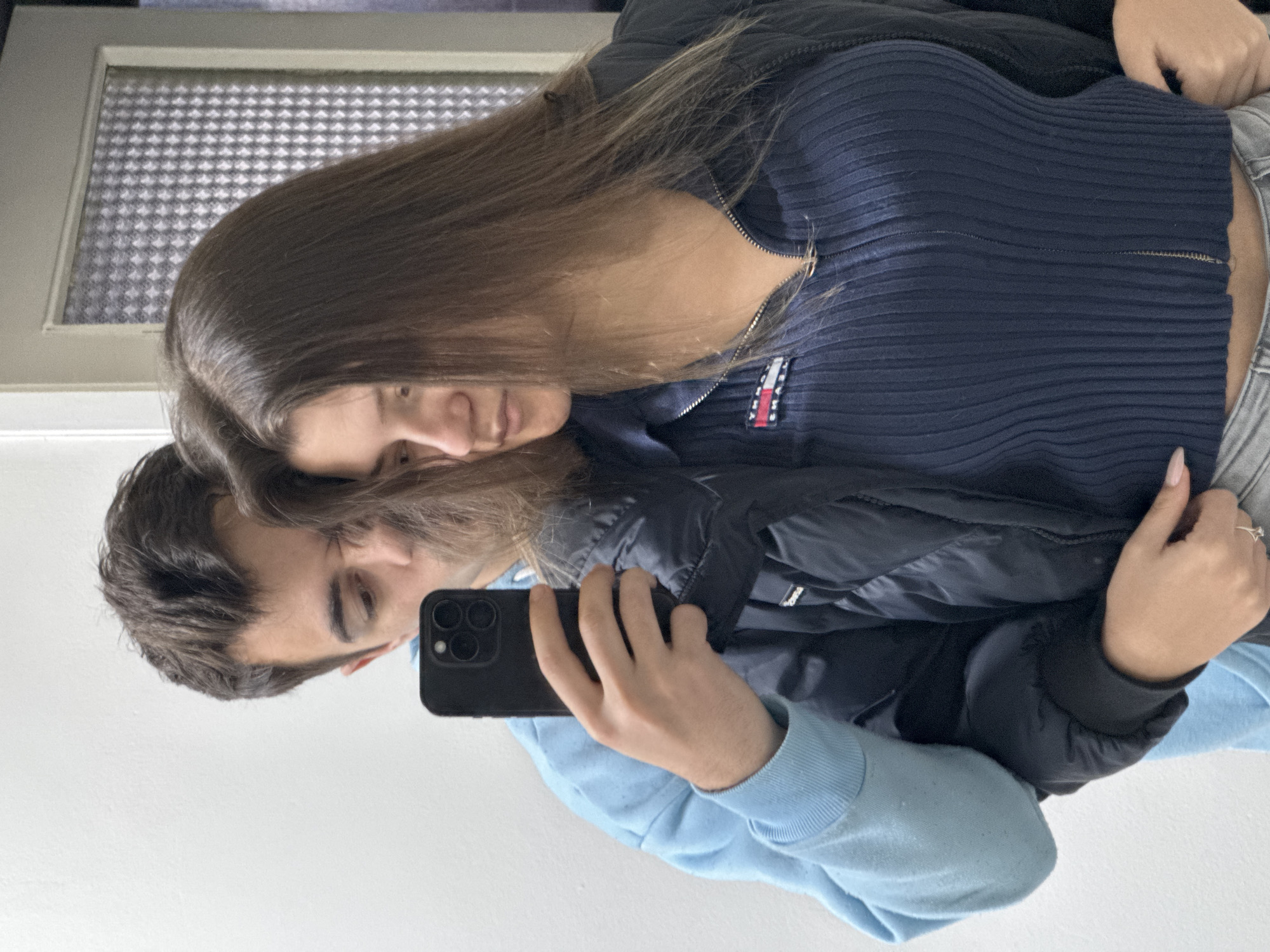A couple is taking a selfie in front of a mirror. The man, with dark hair and wearing a light blue sweater, is holding a black smartphone. The woman, with long brown hair, is wearing a dark blue ribbed top with a small Tommy Hilfiger logo, a black jacket, and grey pants. They are standing very close to each other, with the woman partially behind the man. In the background is a door with a frosted glass panel. The location is likely indoors, but the city of Sofia, Bulgaria, is not evident from the visual details of the image.
