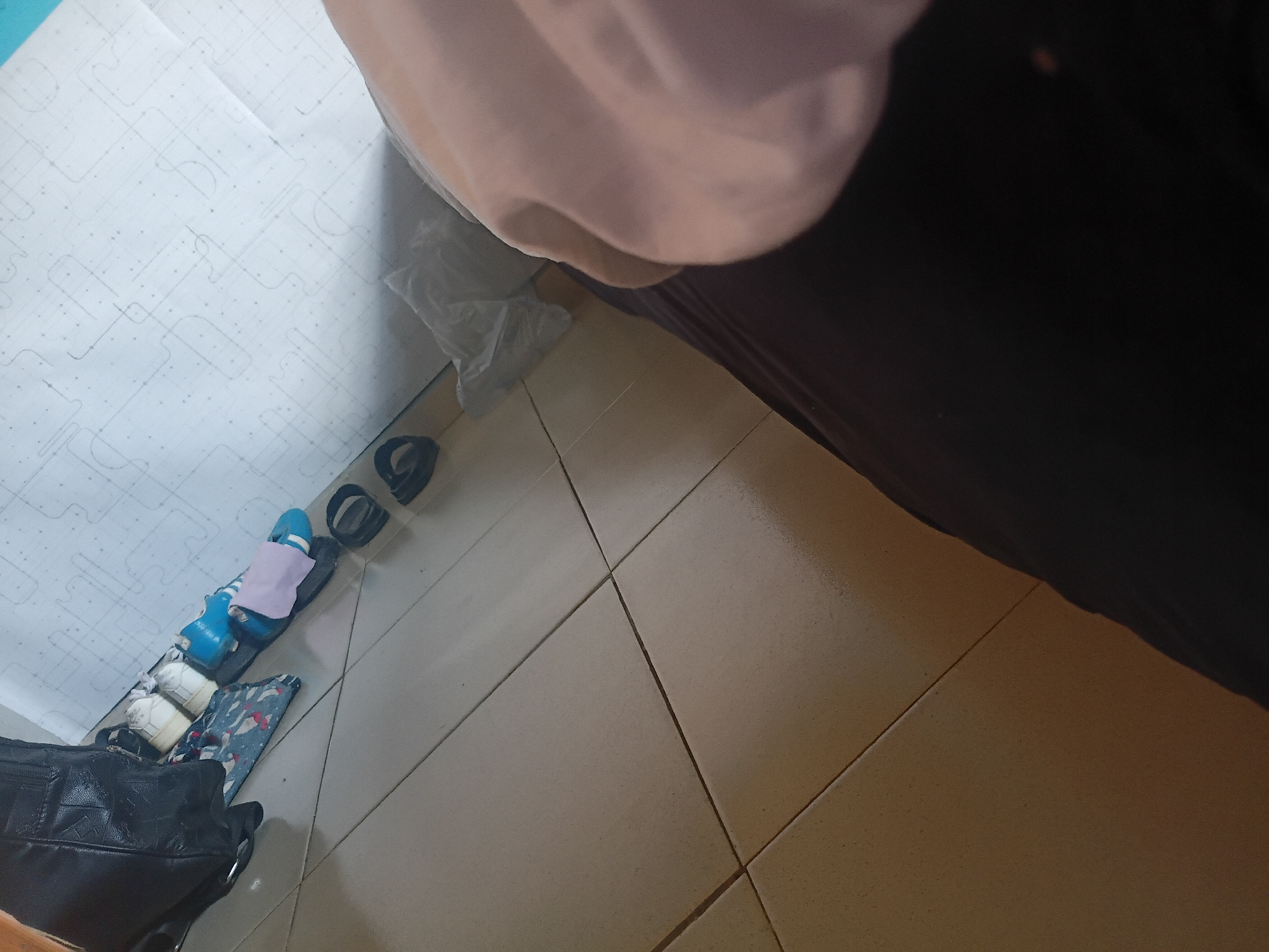 In Jalingo, Nigeria, a scene unfolds on a tiled floor. A black leather boot is positioned to the left, partially obscuring a patterned cloth bag. Next to the bag, a pair of white sneakers and blue sandals with a pink cloth are visible. Further to the right, two black sandals lie on the floor. The background features a white wall with a faint geometric pattern. The lower right portion of the image is dominated by dark fabric, possibly clothing, and a light-colored fabric.