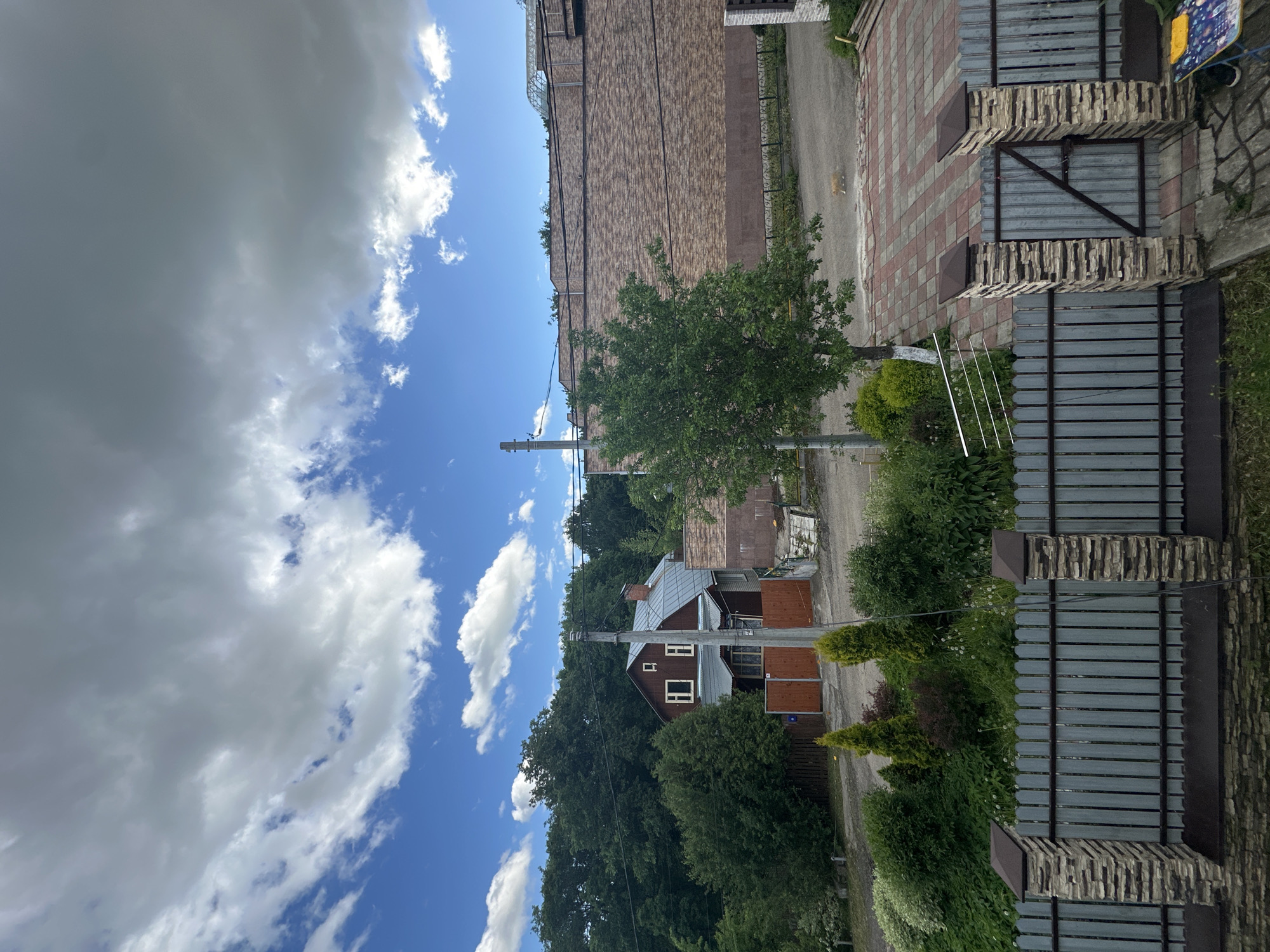 The image depicts a residential scene, likely taken in Nizhny Novgorod, Russia. It shows a two-story house with a brown facade and a gray roof, nestled among green trees. A utility pole stands nearby, with power lines running along the street. Adjacent to the house is a paved road or sidewalk with reddish-brown brick patterns. A tall, brownish brick building is visible in the background. In the foreground, there is a fence made of metal bars and brick pillars. The sky above is partly cloudy with both white and gray clouds scattered against a blue backdrop. There is a light-colored animal, possibly a dog or cat, walking in the road. The picture appears to be taken during the daytime.