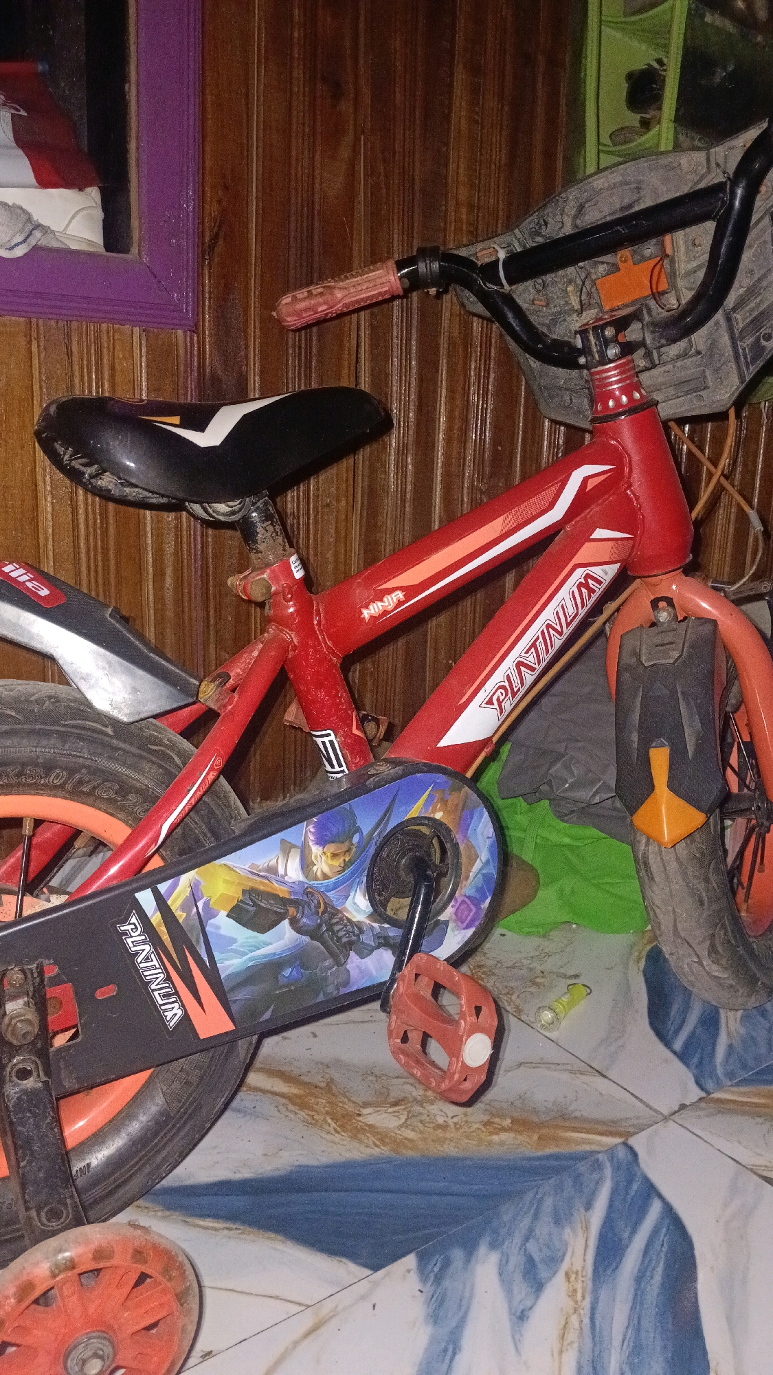 A red children's bicycle is leaning against a wooden paneled wall. The bicycle has a black seat with a white stripe, red handlebars with a pink grip on the left, and a black chain guard with a colorful graphic of animated characters. The front and rear wheels have orange rims. A small red training wheel is visible at the bottom left of the image. A dark gray fender is attached to the rear wheel. The bicycle appears to be indoors, possibly in a residential setting. The floor is tiled with a pattern of blue and brown. A small yellow object, possibly a piece of plastic, is on the floor near the rear wheel. There is no indication of the specific location within Central Jakarta, Indonesia, other than the general description of the objects and environment.