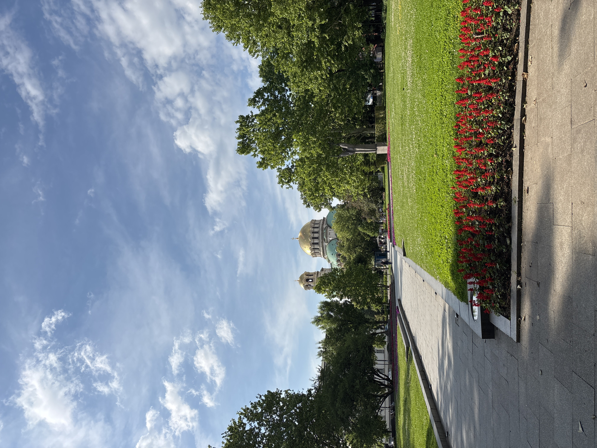 This detailed image depicts the area around the St. Alexander Nevsky Cathedral in Sofia, Bulgaria, viewed from a park or square on a bright, partly cloudy day. The sky occupies the upper quarter of the frame, showcasing a light blue hue with scattered white cumulus and wispy cirrus clouds.
In the foreground, a light grey paved pathway runs along the left side, extending into the mid-ground. Adjacent to the right edge of this pathway is a long, narrow flower bed filled with vibrant red Salvia flowers, arranged in neat rows. To the right of the flower bed, a sprawling, well-maintained green lawn covers a significant portion of the mid-ground, interspersed with small white clover flowers.
In the mid-ground, several mature trees with dense green foliage are prominent, framing the scene. On the right side of the lawn, partially obscured by trees, stands a grey stone statue of a draped figure with arms raised or outstretched, positioned on a low pedestal. Further back on the left, a broader paved area or square is visible, with hints of purple flowers in another distant flower bed. Several indistinct human figures are visible on the pathways and in the distance, appearing to walk or sit.
In the background, the grand St. Alexander Nevsky Cathedral dominates the skyline, positioned slightly left of center. Its distinctive golden domes and smaller green cupolas are clearly visible above the tree line. One prominent bell tower with a golden dome is also visible to the left of the main structure. Parts of other light-colored buildings are discernible behind the trees on the far left. The overall environment suggests a well-kept urban park or memorial area.
