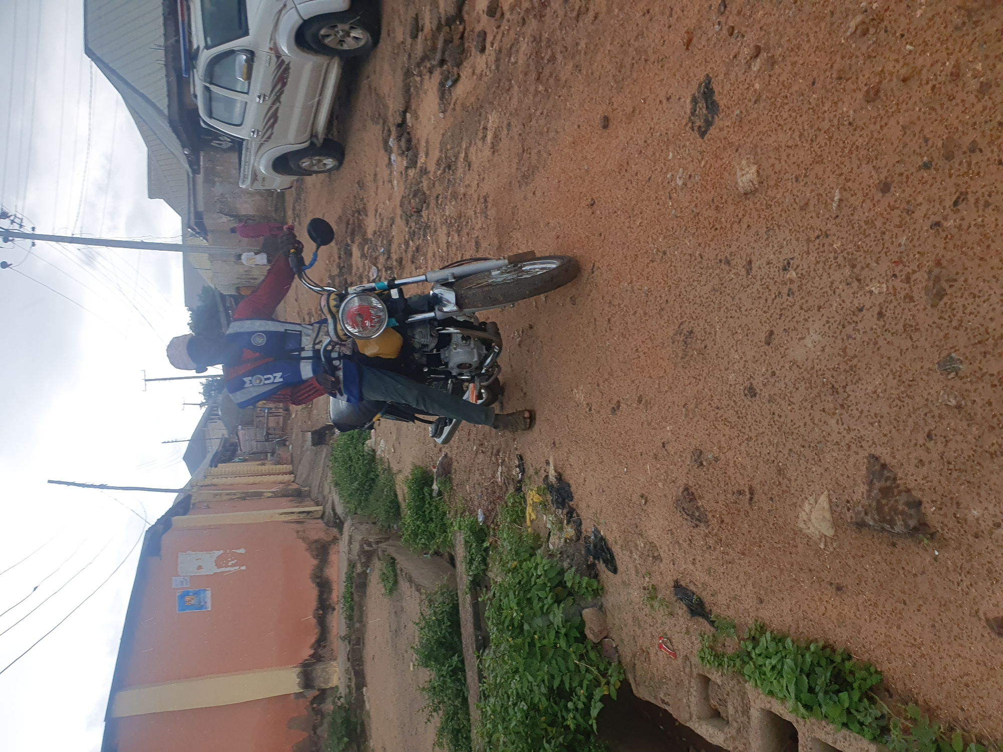 In Lokoja, Nigeria, a person wearing a high-visibility vest over a red shirt and dark pants is seated on a yellow motorcycle. The motorcycle has a chrome exhaust pipe and a circular headlight. The person is wearing sandals and a light-colored cap. The background features buildings with orange and cream-colored walls, some with corrugated metal roofs. A white pickup truck is parked to the right of the motorcycle. The ground is covered in reddish-brown soil, with sparse vegetation and some scattered debris. Overhead, power lines are visible against a cloudy sky. The scene appears to be a street in a residential or commercial area.