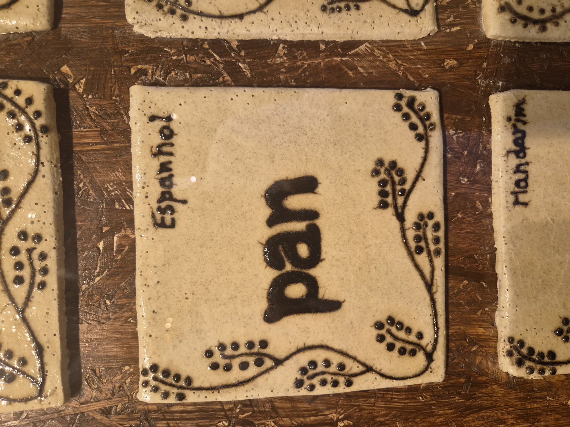The image shows a close-up, eye-level view of four ceramic tiles arranged on a wooden surface. The tiles are rectangular and appear to be decorative elements or part of a larger installation. Each tile is beige with dark brown lettering and vine-like designs featuring small dots.
The top tile displays the word "Espanhol" written in dark brown cursive script. It also has a decorative vine with dots on the left side.
The central tile features the word "pan" in large, bold, dark brown lettering. Similar to the top tile, it has decorative vines with dots on the left and right sides, extending from the edges towards the center.
The bottom left tile has a vine design on its left edge and the word "Mandarim" written in dark brown cursive script.
The bottom right tile is partially visible, showing a dark brown vine design.
The wooden surface beneath the tiles has a rough, textured appearance with visible grain and variations in color, suggesting it might be a tabletop or a wall. The lighting is slightly diffused, highlighting the texture of the tiles and the wooden surface. The image is likely taken indoors.
This image is likely from the Museum of Bread (Museu do Pão) in Seia, Portugal, where similar ceramic tiles are displayed, representing different types of bread in various languages.