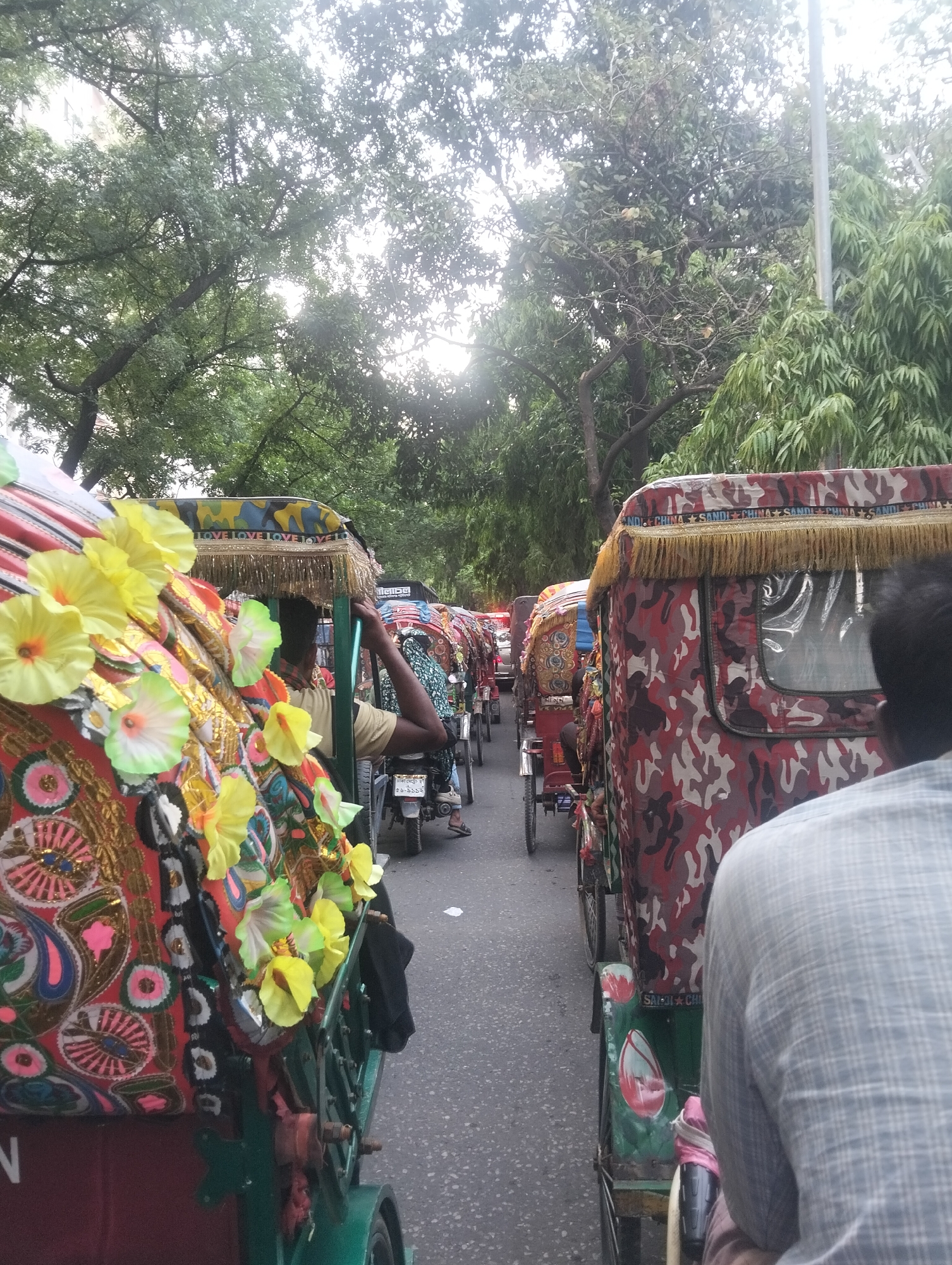 The image shows a street in an unknown city, likely in South Asia, based on the prevalence of cycle rickshaws. The scene is filled with brightly decorated rickshaws in a dense traffic jam on an asphalt road. The rickshaws exhibit a variety of colorful designs, including floral patterns and camouflage prints. Some are adorned with yellow artificial flowers and fringe. There are people seated inside the rickshaws and rickshaw pullers operating the vehicles. The environment is urban, with tall trees providing a canopy above the street. The lighting suggests it is daytime, although partly overcast. The OCR data shows snippets of words and phrases, including "LOVE," "CHINA," and a phone number, potentially identifying the origin or decoration of the rickshaws.
