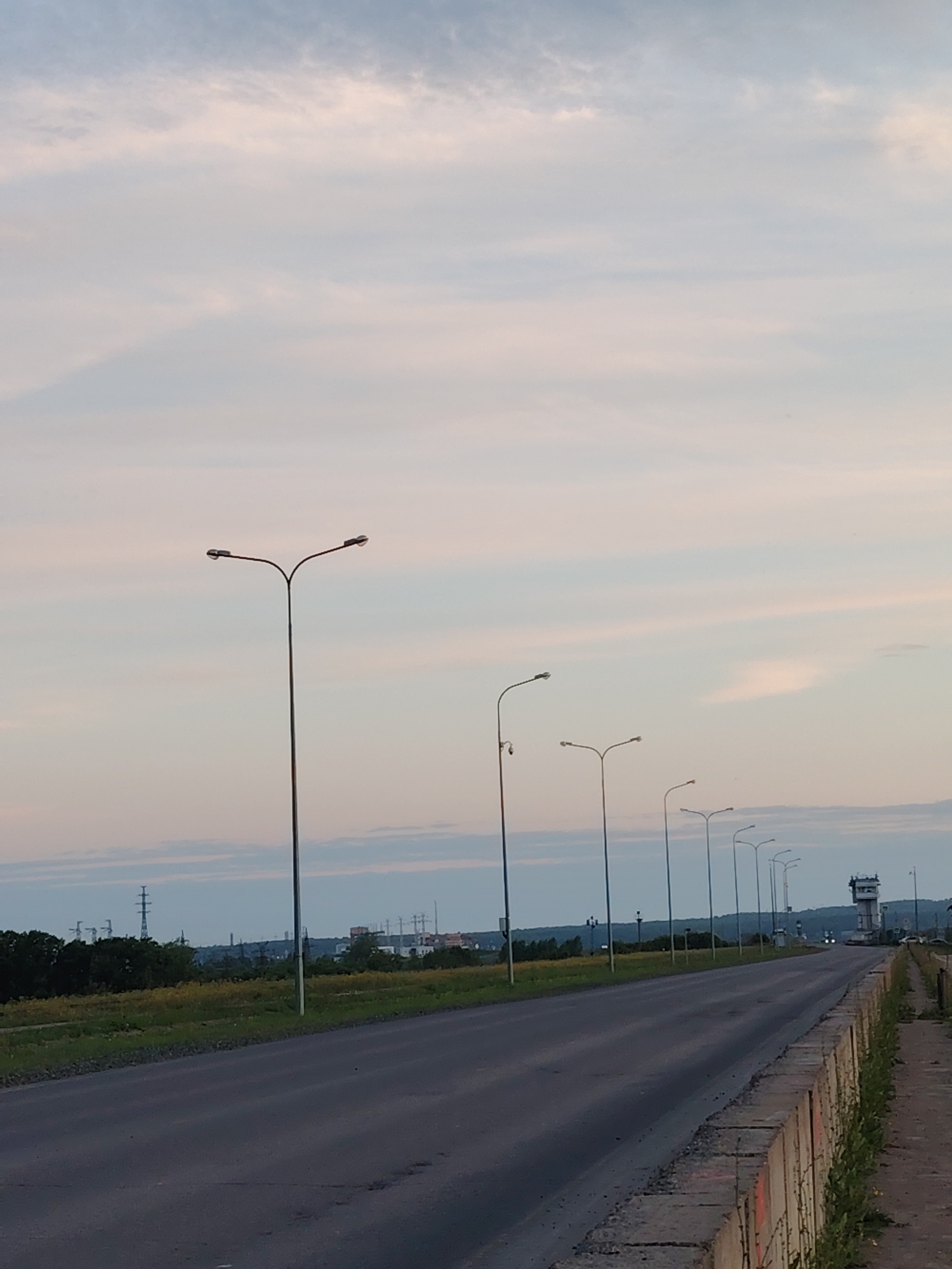 The image shows a road lined with streetlights, stretching into the distance. The sky is overcast with pale clouds. In the background, there are trees and power lines. A tall, narrow building with windows is visible on the right side of the road in the distance. The road appears to be in a semi-urban or industrial area. The perspective suggests the photo was taken from the side of the road, looking down its length. The overall impression is one of a clear, twilight sky over a somewhat desolate landscape. The scene is devoid of people.