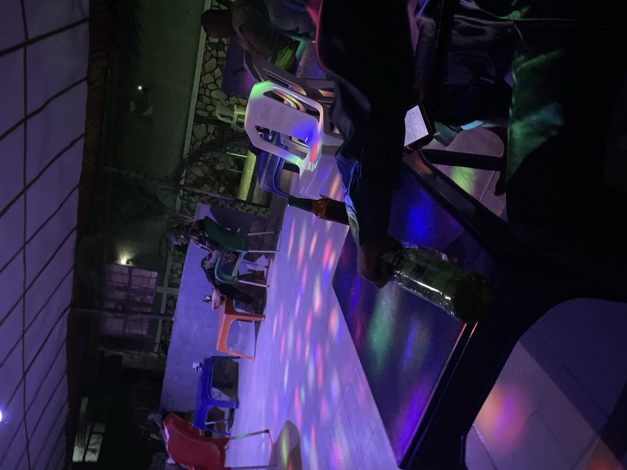 In Madalla, Nigeria, an outdoor establishment with a covered roof and ambient lighting is depicted. The scene includes multiple individuals seated and interacting within the space. A prominent white plastic chair is visible with "God Bless You" inscribed on its backrest. The floor is illuminated by colorful lights, suggesting a festive atmosphere. Several tables and chairs are present, some occupied by people and drinks. One person is holding a green glass bottle, and another is looking at a mobile phone. The background shows a building with windows and stone-like wall decorations. The overall impression is of a social gathering at night.