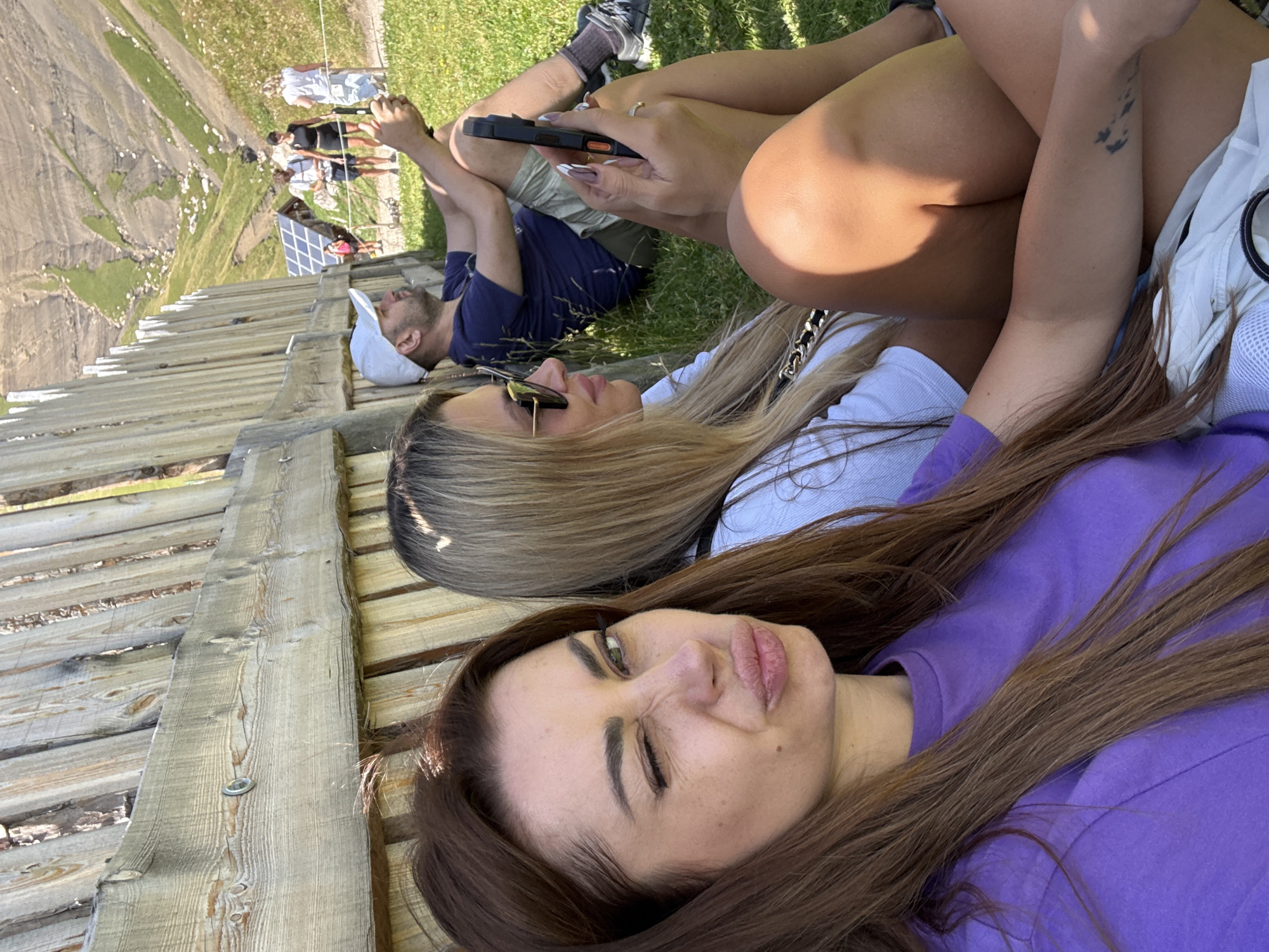 The image shows a group of people resting on a grassy slope near a wooden fence. In the foreground, a woman with long brown hair is taking a selfie, winking at the camera with her lips puckered. She is wearing a purple t-shirt. To her right, a woman with blonde hair and sunglasses is looking at her phone. Behind them, a man with a white cap is sitting and looking away from the camera. Further in the background, other people are visible walking on a path, with solar panels and a building in the distance. The background features a mountainous landscape with green slopes. The setting appears to be an outdoor recreation area, possibly in the Italian Dolomites, given the mention of Campitello di Fassa, Italy.