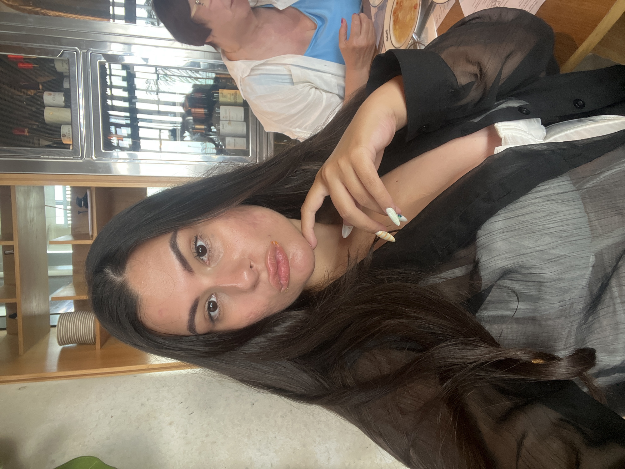 The image presents a woman with dark hair, a fair complexion, and defined facial features, taking a selfie. She is wearing a black, semi-transparent garment, layered over a white top. Her hand is near her face, displaying long nails. The background shows elements of a restaurant setting, including a refrigerated display case containing bottles, presumably wine. Another person is visible in the background. The location can be identified as Tel-Aviv, Israel by the "Tel-Aviv, Israel" printed on the label of a wine bottle visible in the background.