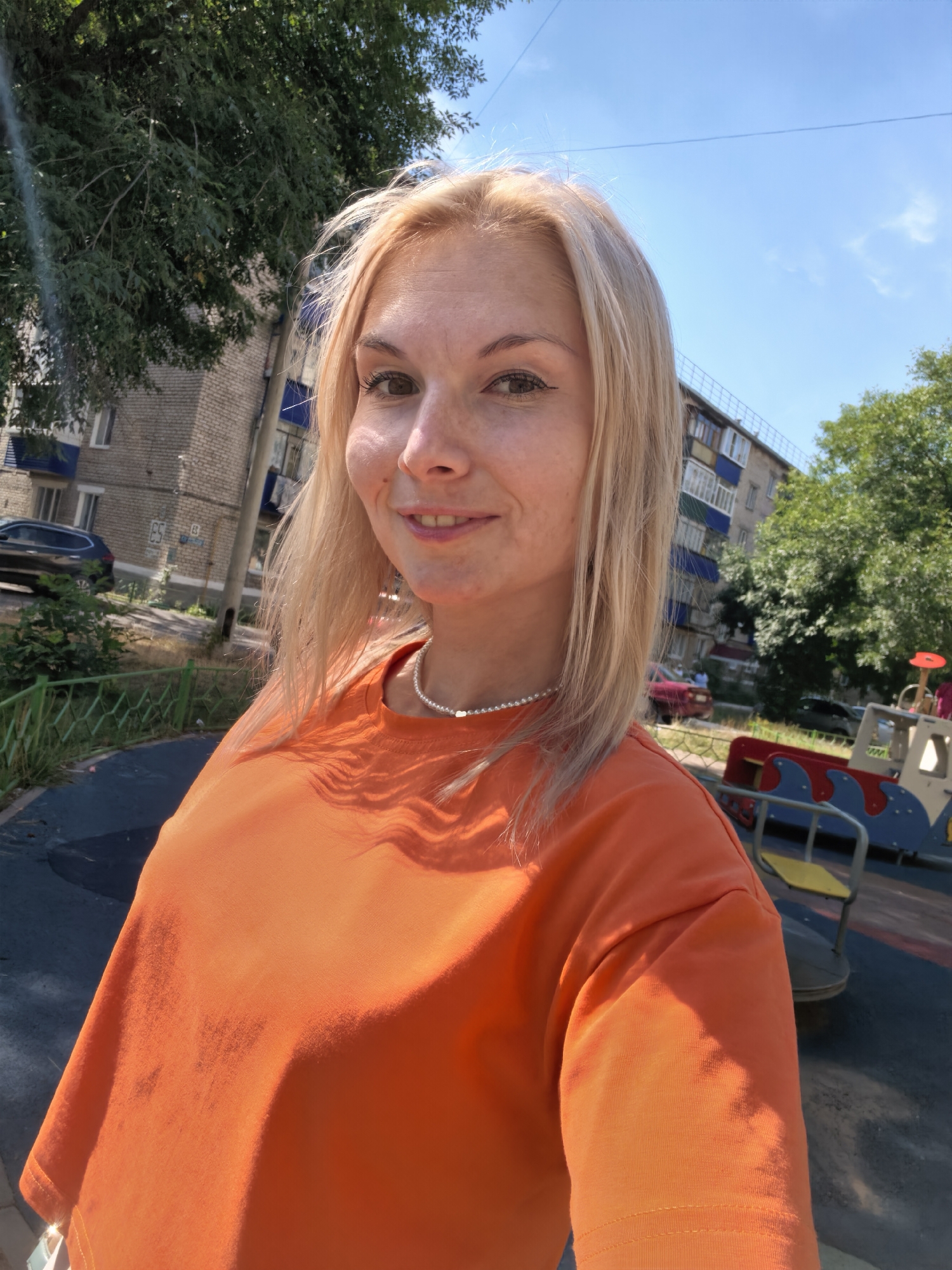 A young woman with blonde hair and light skin is captured in a selfie outdoors. She is wearing an orange t-shirt and a pearl necklace. The background suggests a playground area with colorful equipment, and beyond that, an apartment building and trees are visible. The sky is blue with some scattered clouds, indicating a sunny day. The woman is smiling at the camera. The scene appears to be in a residential area within the city of Sterlitamak, Russia, given the architectural style of the building and the general urban environment.