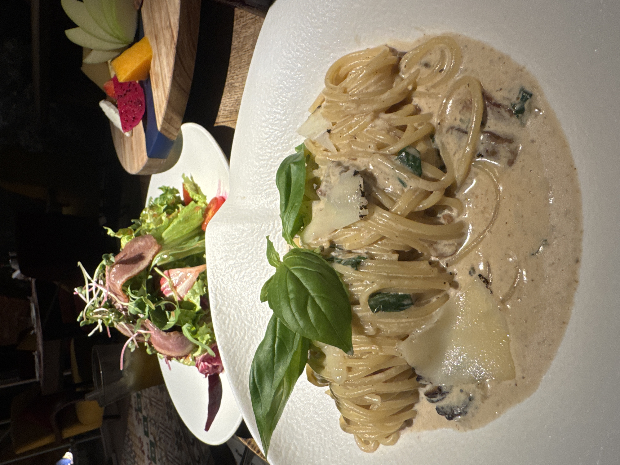 The image shows a close-up, overhead view of a meal served at a restaurant in Hanoi, Vietnam. The main subject is a white, textured plate filled with spaghetti in a creamy sauce. The pasta is intertwined and topped with shavings of Parmesan cheese and fresh green basil leaves. Small pieces of what appear to be mushrooms and herbs are visible within the sauce.
To the left of the pasta, a smaller white plate holds a vibrant salad. The salad consists of fresh green lettuce, microgreens, thinly sliced red radishes, and strips of cured meat, possibly ham or prosciutto. A single red cherry tomato is also visible.
In the background, a wooden board displays a selection of sliced fruits, including green apple, mango, and dragon fruit. The lighting suggests it is nighttime or the restaurant is dimly lit. The overall scene depicts a well-presented meal, hinting at a dining experience. There is no visible text in the image.