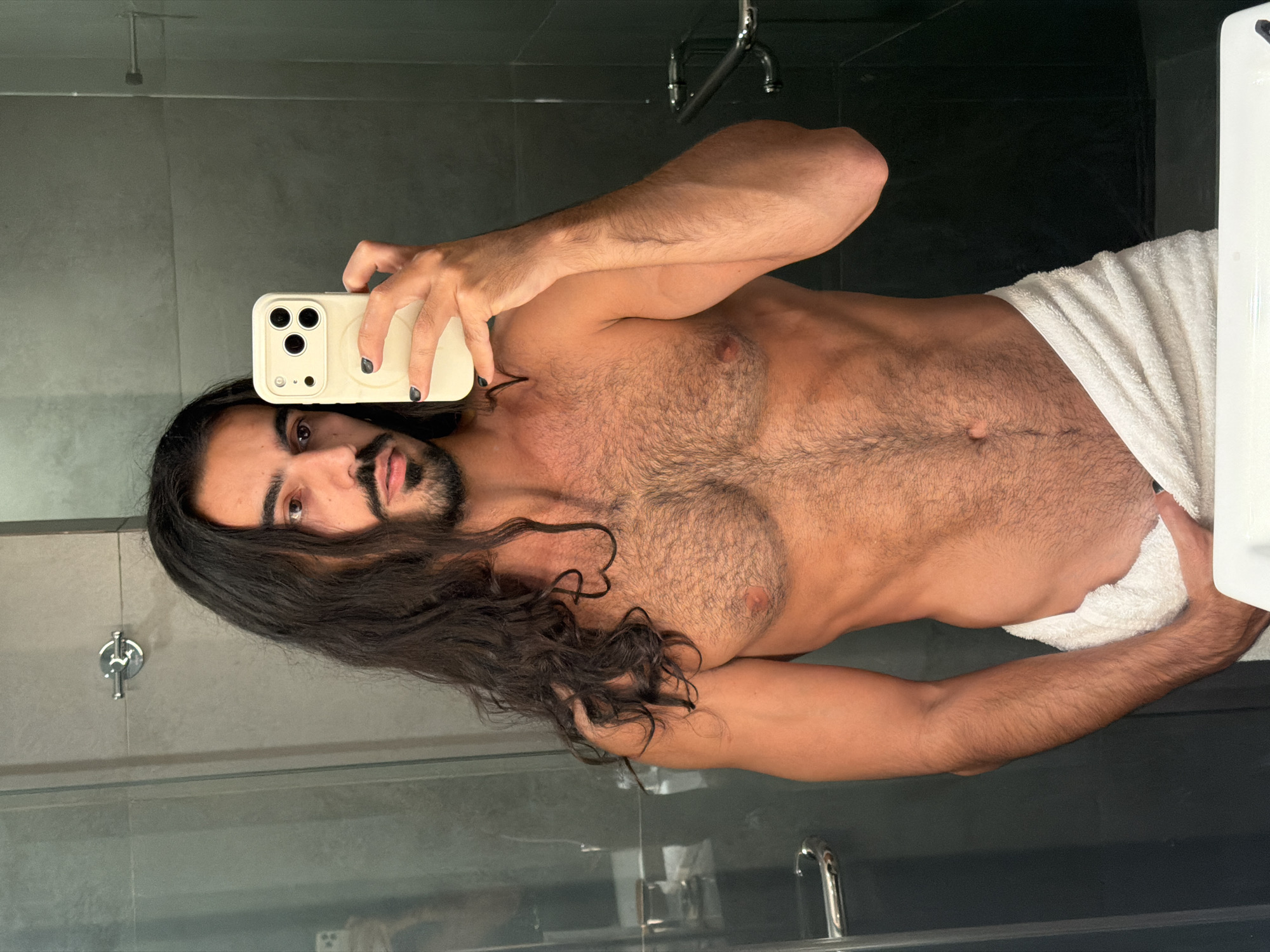 The image is a mirror selfie taken in a bathroom. A person with long dark hair, a beard, and mustache is holding a smartphone in their right hand, taking the picture. They are wearing a white towel around their waist. The bathroom has dark tiled walls and a silver faucet is visible on the wall. There are also silver handles on a glass door. The person's chest and abdomen are exposed and covered in dark hair. The person has dark nail polish on their fingernails. The reflection of a sink is visible in the foreground. The location is not specified in the image, and there is no information about Dadapan, Indonesia.