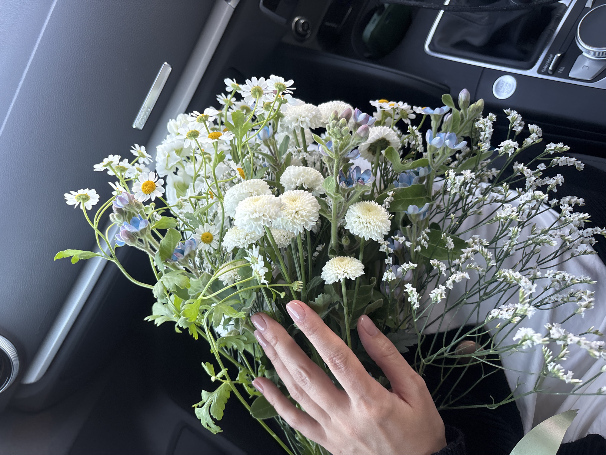 A bouquet of white and blue flowers rests in the passenger seat of a car. A female hand with manicured nails gently touches the flowers. The interior of the car is visible, including the dashboard, console, and start/stop button. The location is indeterminate.