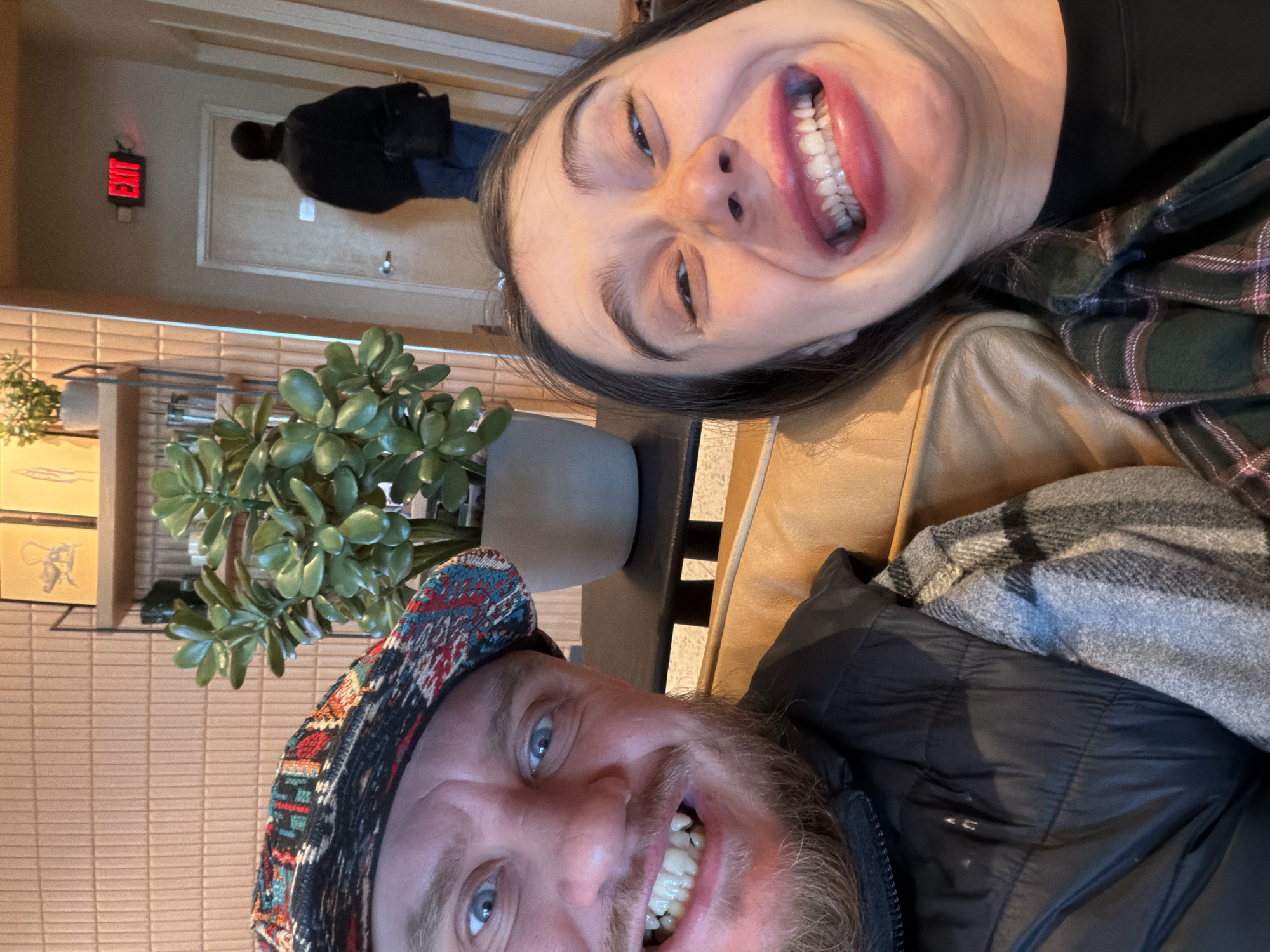 The image depicts two individuals posing close together, likely for a selfie. The person on the left, positioned at the bottom of the frame, is a bearded man wearing a patterned cap and a dark jacket. He is smiling broadly. To the right and at the top of the frame, a woman is also smiling widely, showing her teeth. She has dark hair and is wearing a patterned shirt. The background includes a potted plant, a tan-colored wall, and what appears to be an exit sign and a person walking through a doorway in the distance. This suggests the location is indoors, possibly a cafe or restaurant in Morton Grove, United States of America.