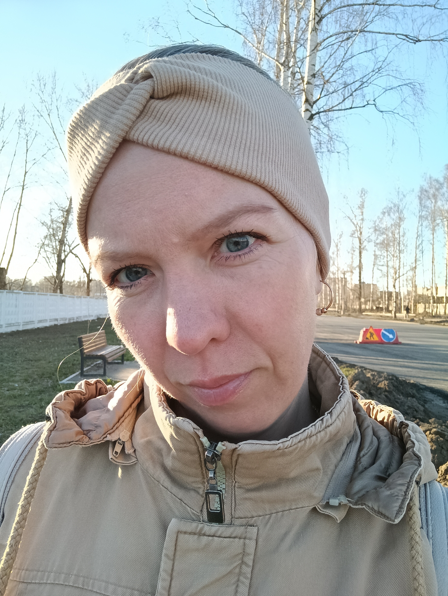 A medium shot captures a woman in Chaikovsky, Russia. She has fair skin, blue eyes, and wears a beige, twisted knit headband and a matching tan hooded jacket. The jacket is partially zipped up and features a braided cord on the side. She is outside in a city environment. Behind her, there is a road with a road sign indicating road work. Beyond the road, buildings and bare-branched trees are visible under a clear blue sky. A park bench can be seen to the left. The lighting suggests it is daytime.