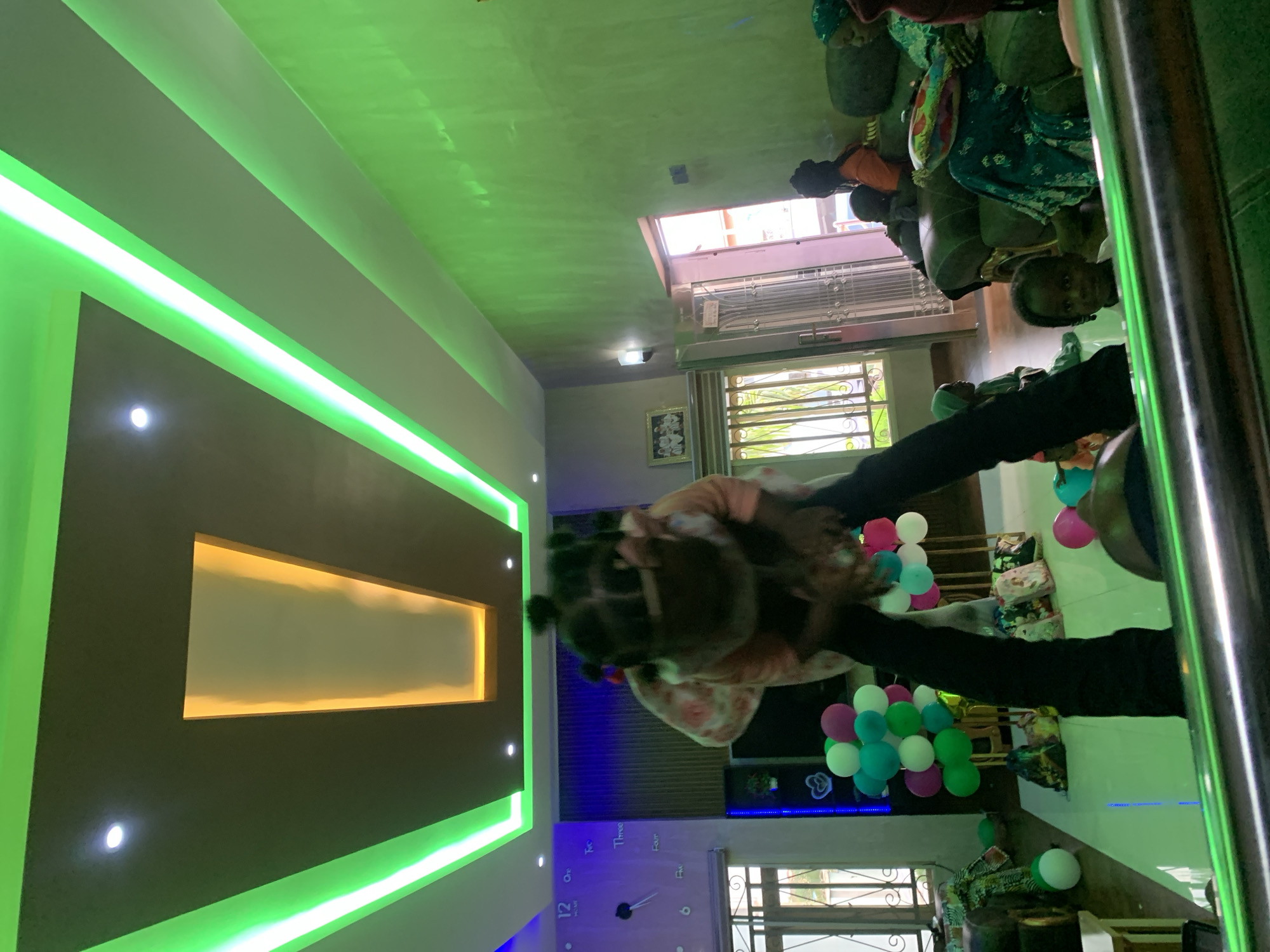 The image captures an indoor scene, likely a living room or event space, in the city of Jos, Nigeria. The room is characterized by contemporary design elements, including a multi-layered ceiling with distinct lighting. The central ceiling features a dark brown/grey panel with a recessed rectangular section illuminated by a warm yellow-orange light. White panels flank this central area, bordered by bright green LED strip lights that cast a strong green hue throughout the upper part of the room, particularly on the right wall. Several white recessed spotlights are also present on the ceiling.
In the center of the foreground, a young child with dark, braided hair is being held aloft by an obscured adult. The child, wearing a light-colored top with a floral pattern and dark pants, is looking downwards with her arms extended. On the light-colored, reflective floor beneath them, several clusters of balloons in various colors including light green, white, pink, and purple are arranged, suggesting a celebratory occasion. Decorative bags or small packages are also visible on the floor.
To the left, a large, minimalist wall clock displaying numbers "12" through "5" is mounted on a textured purplish-blue wall. Below the clock, a window with ornate metal grilles is visible, allowing natural light into the room. Adjacent to the window, a dark shelf unit or entertainment stand with blue accent lighting holds a few small items.
The right side of the room is dominated by a green-painted wall, strongly illuminated by the green ceiling lights. A doorway with an open light-colored door reveals a brightly lit area beyond, possibly an exterior space or hallway, with orange-red tones visible. A wall-mounted light fixture is positioned near the doorway. Several individuals are seated on dark sofas in the midground and background. An adult female wearing a green headscarf and a patterned dress is seated on a sofa on the far right. Another child with dark, braided hair, wearing a light-colored top, is visible in the lower right foreground, looking directly towards the camera, partially obscured by a dark horizontal bar. Other individuals, including children and adults, are partially visible seated on the sofas, observing the scene.