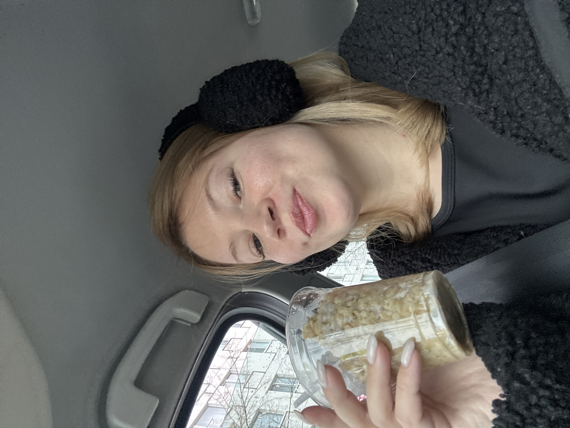 A woman is in a car, holding a clear plastic container of what appears to be oatmeal or porridge. She's wearing black earmuffs and a dark, possibly fleece jacket. The scene is framed by the car's interior, including the ceiling, door handle, and window. Outside the window, the facade of a multi-story building and bare tree branches are visible. Given the style of architecture visible through the window, and the instructions to include the location, it is likely that the location is the city of Chaikovsky, Russia.