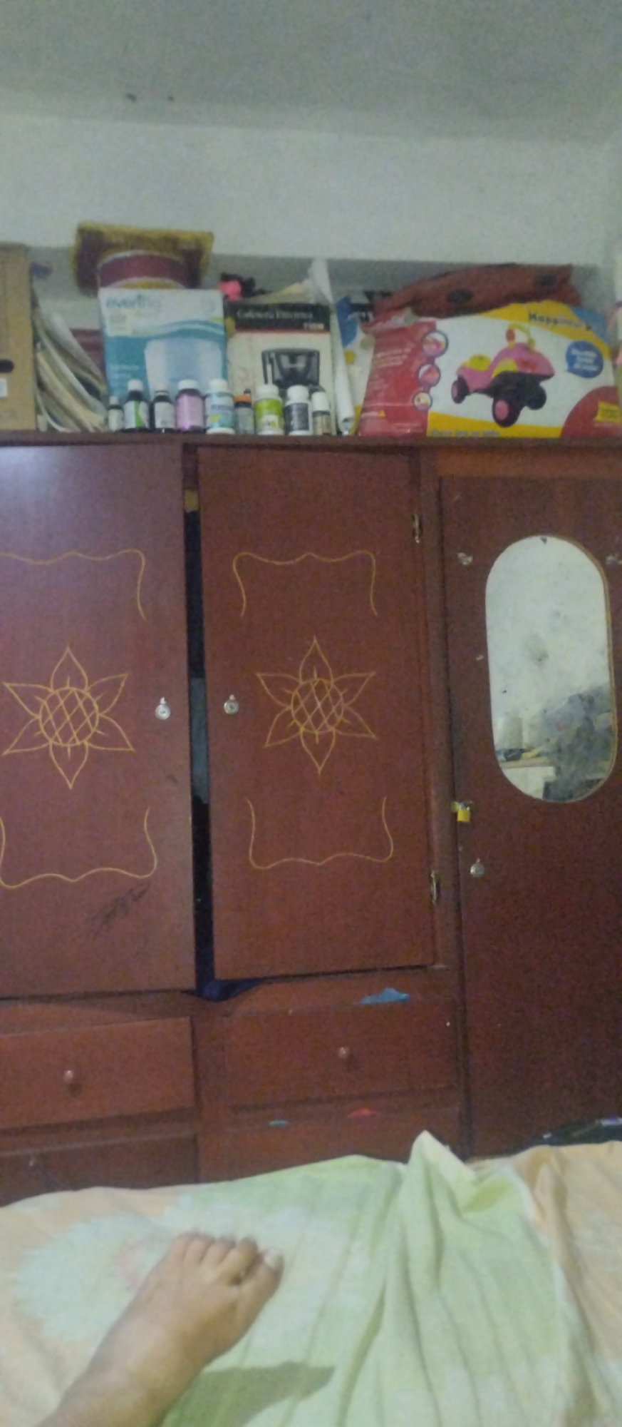A photograph taken indoors shows a person's left foot on a bed in the foreground, with a wooden wardrobe taking up the majority of the background. The wardrobe is comprised of three sections: two doors with decorative, sunflower-like designs on the left and center, and a third door on the right with an oval mirror. Above the wardrobe is a shelf with miscellaneous items, including bottles, boxes, and a child's toy box. The image quality is somewhat blurry. The location is Altagracia, Venezuela.