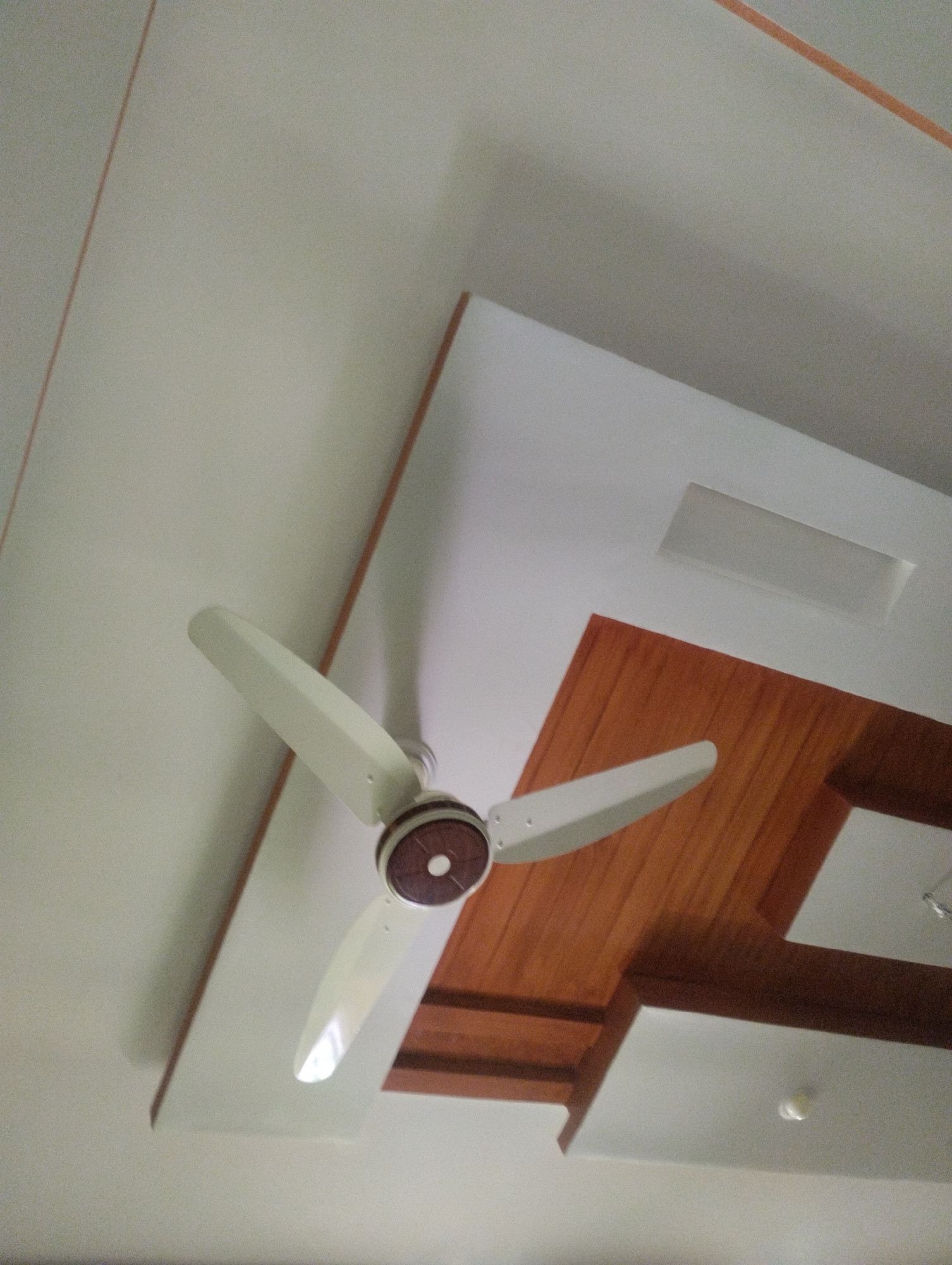 An overhead shot shows a white ceiling fan with three white blades. The fan is mounted on a white ceiling with some textured areas and wooden panels. The wooden panels are rectangular and arranged in a way that suggests a recessed lighting fixture. The ceiling has thin, reddish-brown lines running diagonally across it. The scene is indoors and well-lit.