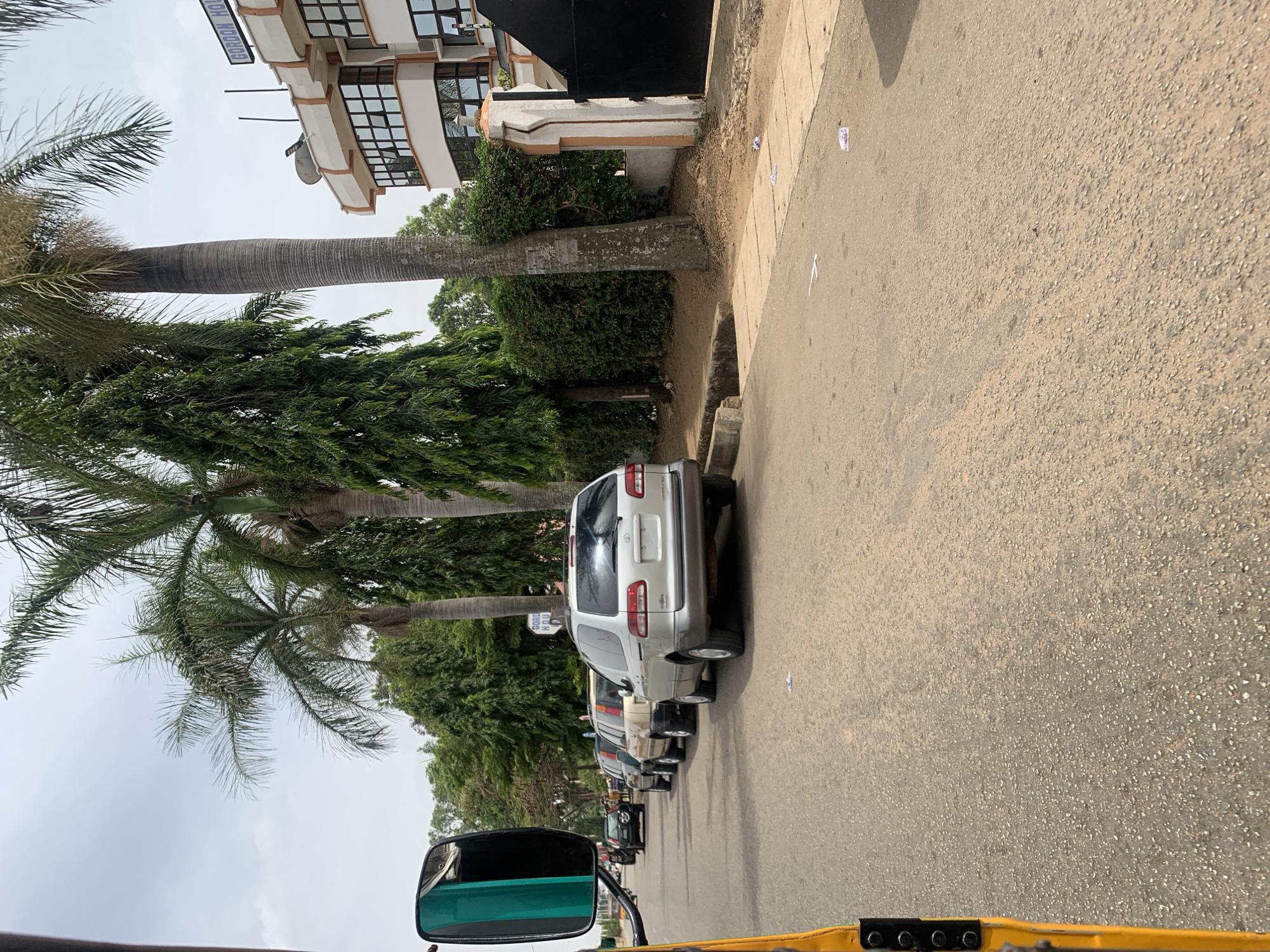The image shows a street scene in the vicinity of New Stadium, Nigeria. A line of parked cars stretches along the right side of the road. The nearest car is a silver SUV. Palm trees and other greenery line the side of the road. A building is visible behind the trees. The photograph appears to have been taken from inside a vehicle, with part of the vehicle's interior visible in the foreground. The sky is overcast.