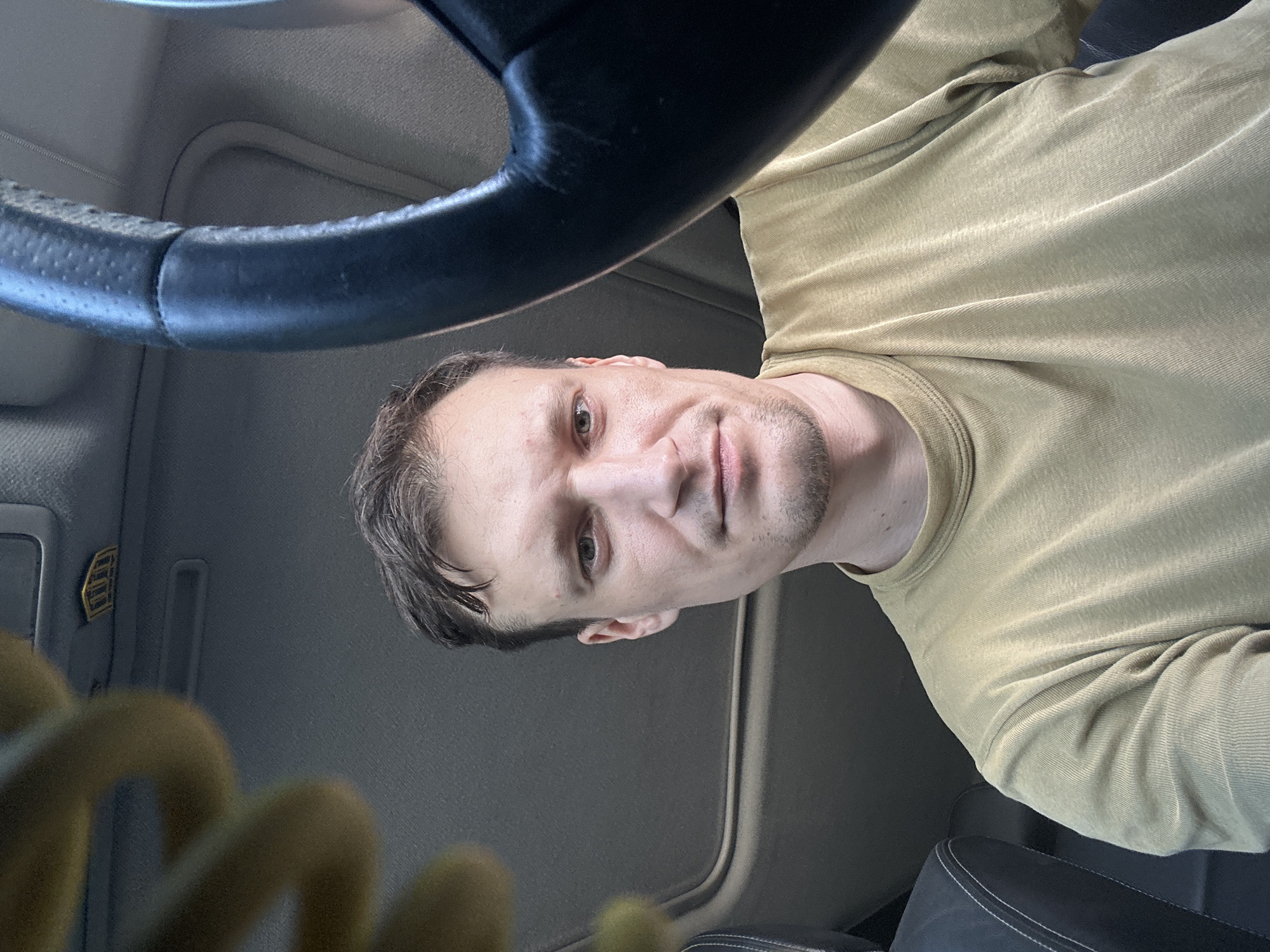 A medium shot, eye-level selfie taken from inside a car, captures a man in his early 30s with short brown hair and a light stubble. He is wearing a tan crew-neck t-shirt. The man is looking directly at the camera. To the right of the frame, a portion of a black leather steering wheel is visible. The car's interior is dimly lit, with a sunroof open overhead, allowing natural light to filter in. A yellow object, possibly part of a car accessory or a toy, is partially visible in the upper left corner. The scene appears to be a typical car interior, with the focus on the driver. The location is not explicitly indicated within the image but is stated to be Shatava, Ukraine.