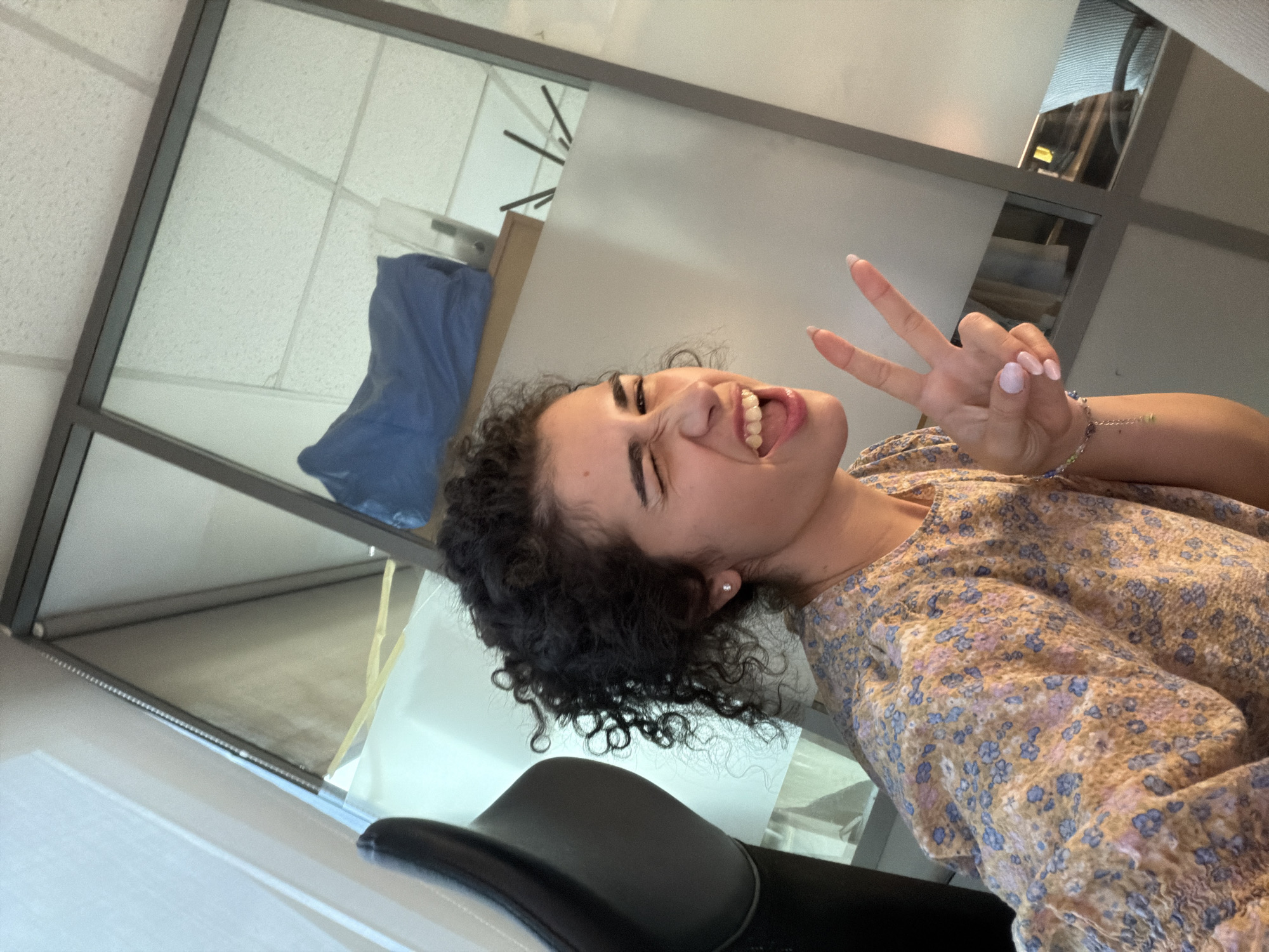 A young woman with curly dark hair smiles widely and makes a peace sign with her right hand. She is wearing a floral print dress with blue and purple flowers on a light-colored background. Her nails are painted a light purple color. A bracelet with beads is visible on her wrist. The image appears to be a selfie taken indoors, possibly in an office or classroom setting, given the presence of office ceiling tiles and glass partitions. A blue tarp is visible through the glass partition in the background. The lighting suggests it could be daytime. The woman's expression conveys a sense of joy and playfulness. The location context provided is Sofia, Bulgaria.