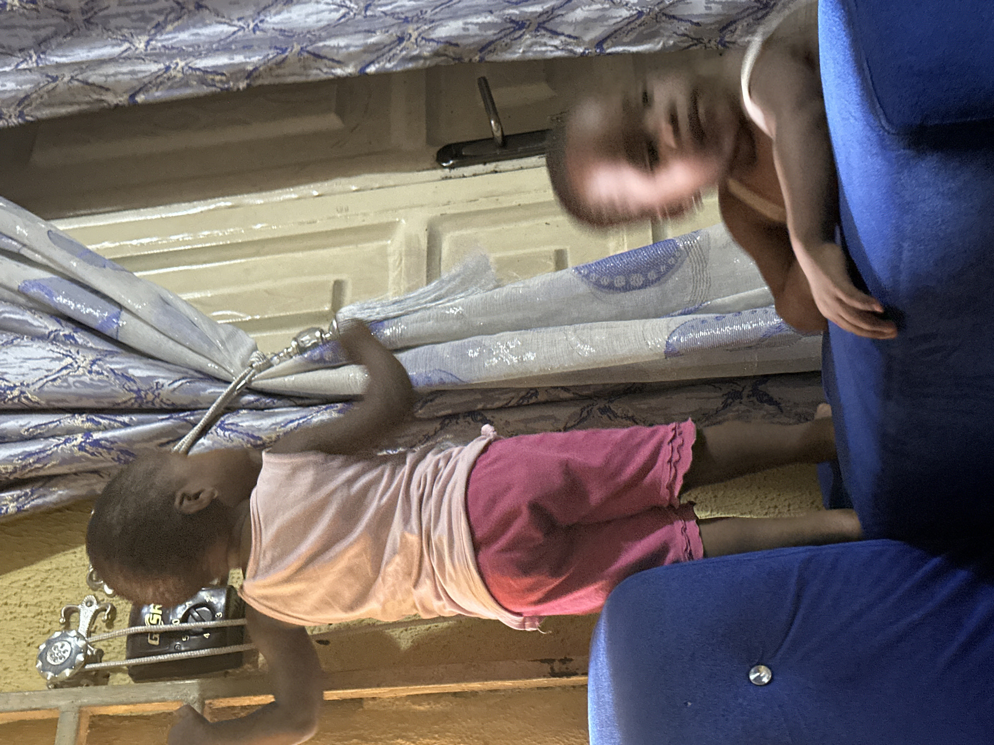In Narayi, Nigeria, a young child, likely a toddler, stands on a blue sofa. The child, wearing a pink tank top and pink shorts, is positioned with their back partially to the camera, facing a wall with a window and curtains. To the left of the child, a black electrical box with a dial and numbers is mounted on the wall. The curtains are patterned with blue and white designs. In the foreground, another child, partially obscured and out of focus, rests their arm on the back of the same blue sofa. The scene is indoors, with artificial lighting casting shadows.