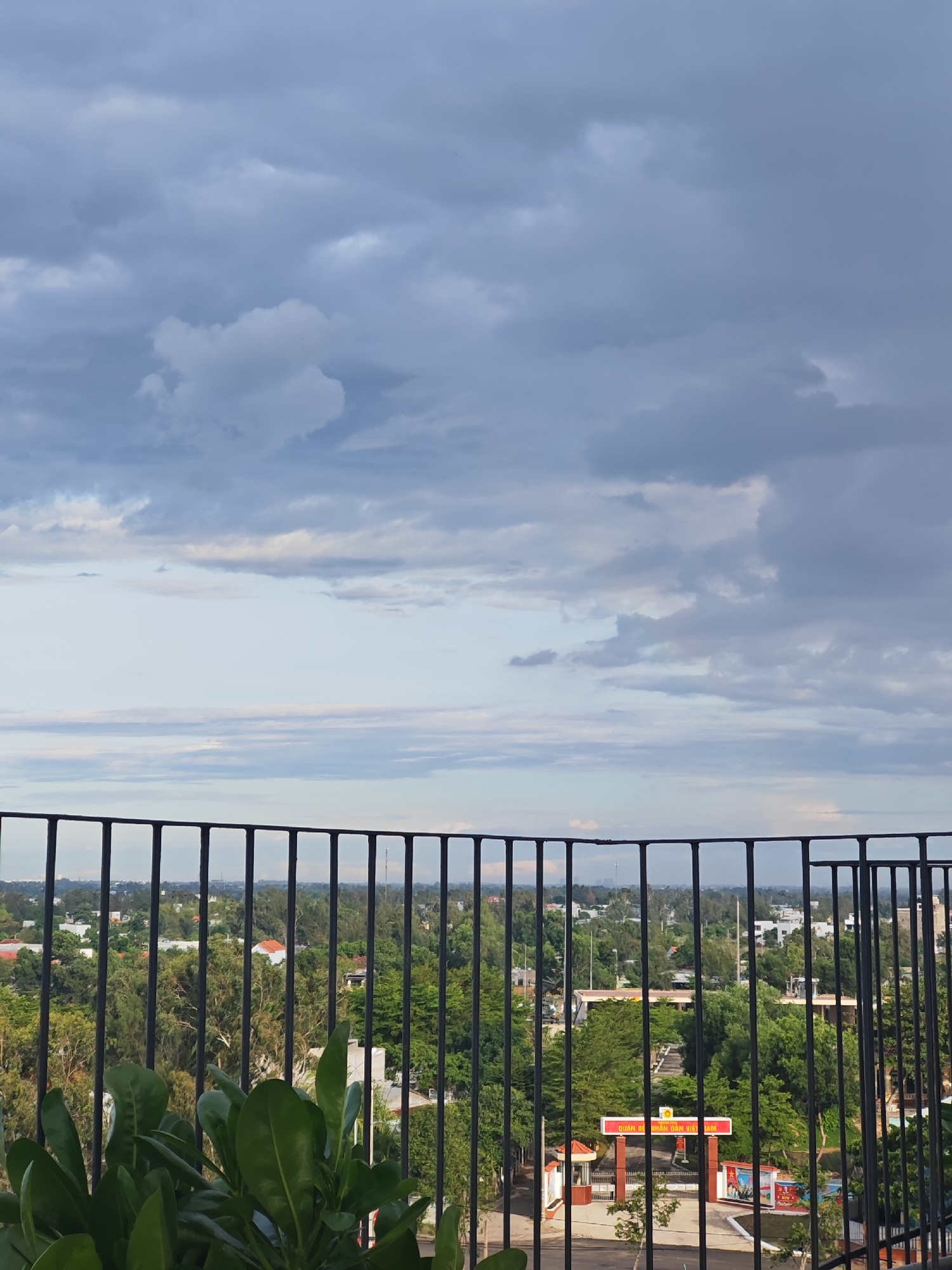 The image is a high-angle shot of an urban landscape in Da Nang, Vietnam, viewed from behind a black metal railing. The sky is filled with dramatic, grey, and white clouds, suggesting impending rain or overcast weather. Below the sky, a hazy horizon displays a mix of buildings, trees, and vegetation. The foreground is partially obscured by lush green foliage, also seen behind the railing. A prominent red and yellow sign with Vietnamese text and imagery is visible in the mid-ground, appearing to be an entrance to a public facility or establishment. The overall scene depicts a typical urban environment in Da Nang, characterized by a blend of natural elements and man-made structures under a brooding sky.
