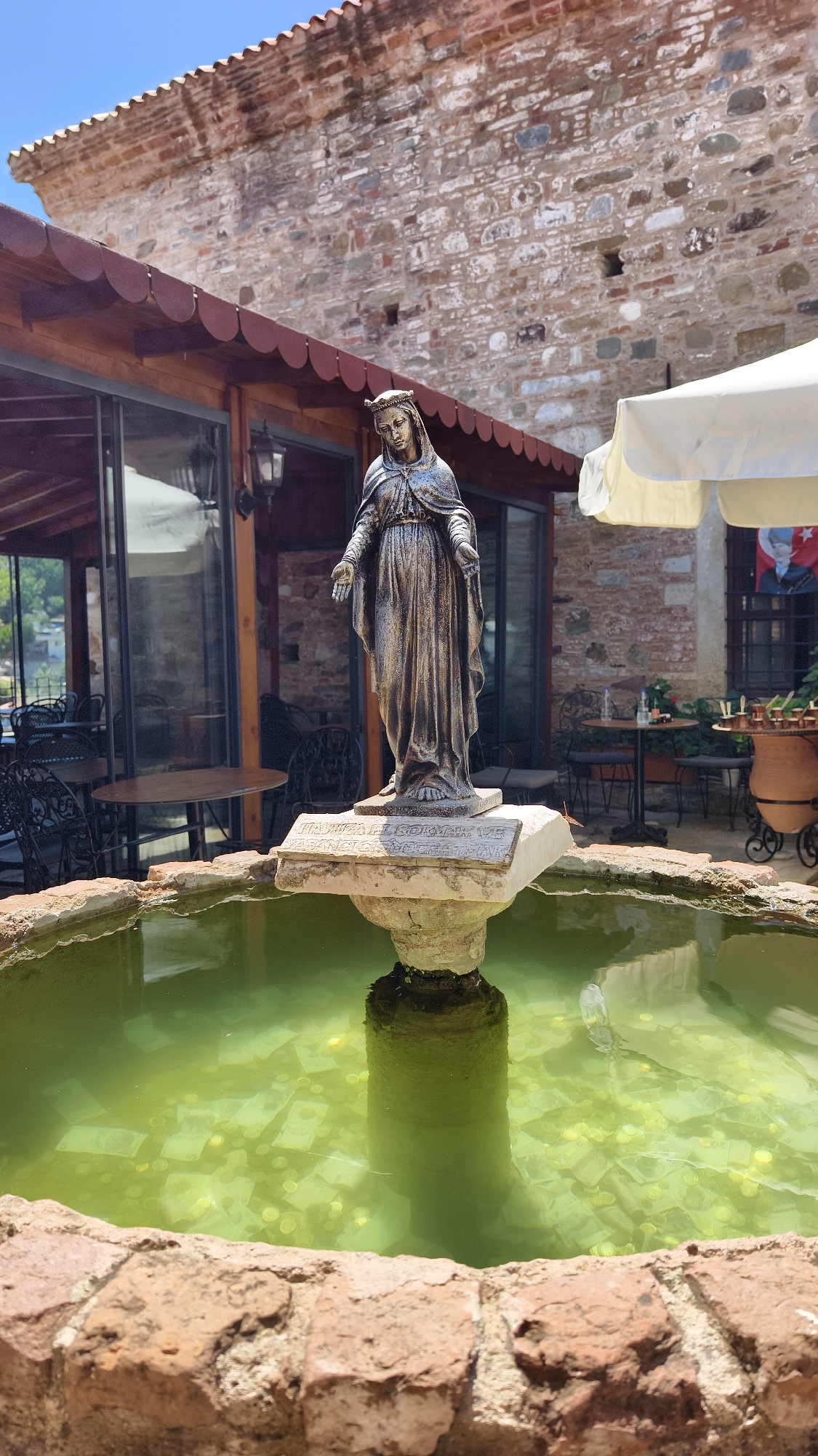 A statue of the Virgin Mary stands in the center of a fountain filled with green water, coins, and paper money. The statue is made of metal and depicts Mary with her arms outstretched, wearing a crown and a long robe. The fountain is made of brick and is surrounded by a tiled patio. In the background, there is a building with a stone facade and a red-tiled roof, and outdoor seating with tables and chairs. The overall scene suggests a religious or historical site, possibly a place of pilgrimage. The location is the House of the Virgin Mary in Selçuk, Turkey.