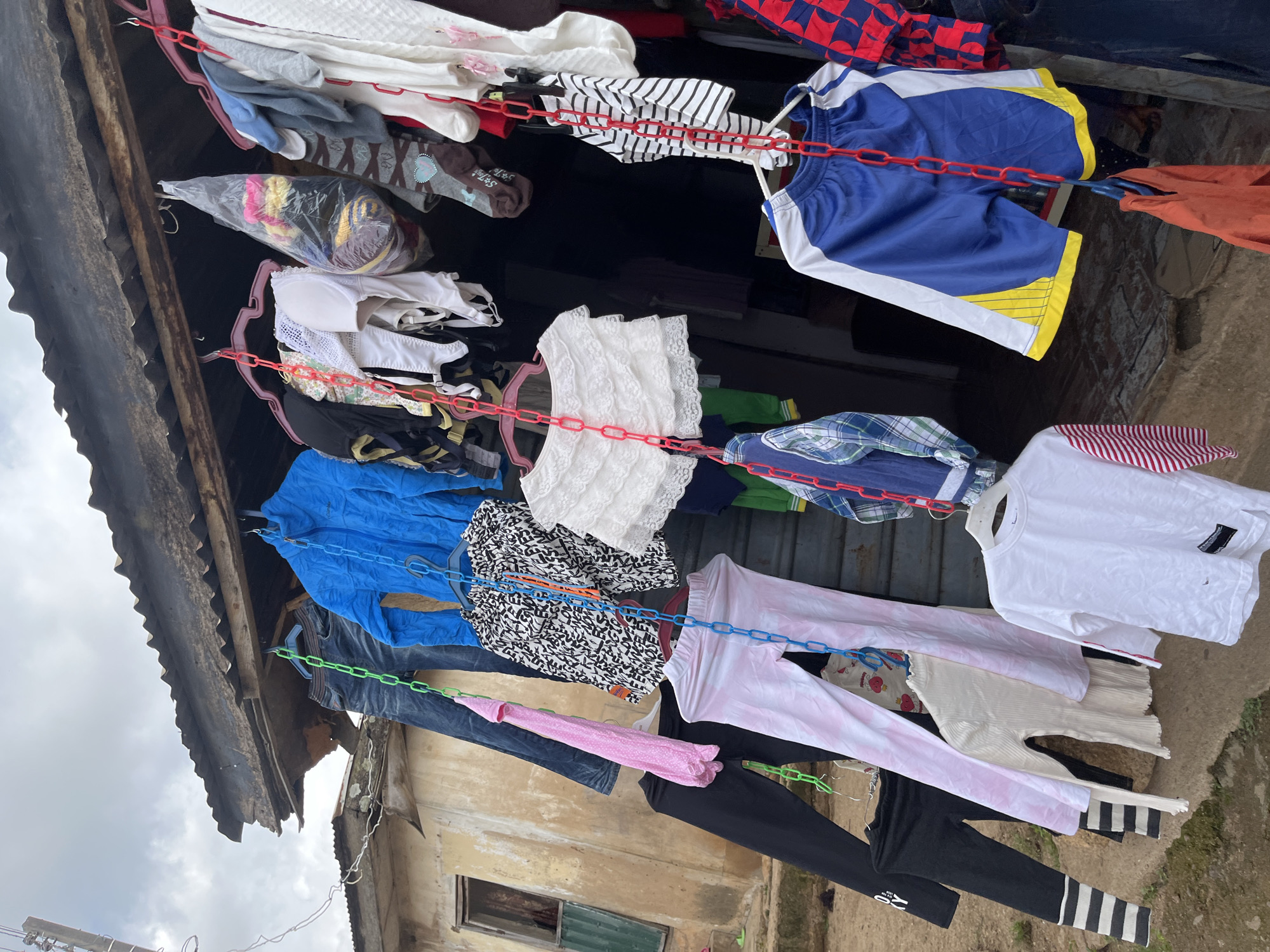 The image depicts an outdoor clothesline in Jos, Nigeria, with laundry hanging to dry. The scene is dominated by various items of clothing, including shirts, shorts, pants, and what appear to be undergarments. The clothes are suspended from metal chains and plastic hangers, suggesting a practical and possibly improvised drying setup.
The setting appears to be a residential area, with parts of a building visible in the background. The building's facade is weathered, showing signs of age and possibly some disrepair, with a corrugated metal roof partially covering the clothesline. The ground beneath the clothesline is rough and uneven, with dirt and debris.
The lighting suggests it is daytime, with natural light illuminating the scene, though the sky is overcast, indicating potentially cloudy weather. There are no people visible in the frame.
Notable details include the variety of clothing, from casual wear like shorts and t-shirts to what might be more formal items. The use of brightly colored chains (red and blue) adds a touch of vibrancy to the otherwise utilitarian scene. There is a plastic-wrapped bundle of clothes hanging, possibly for protection or storage. Some of the clothing has patterns, such as stripes and a black and white print with what looks like abstract lettering.
One piece of clothing, a white t-shirt, has a small black tag with white text, but it is illegible. Another item of clothing, black leggings with white stripes at the ankle, has a white logo with black text.
Overall, the image captures a common sight in many parts of the world, particularly in regions where outdoor clotheslines are prevalent for drying laundry. The scene conveys a sense of everyday life and domestic activity.