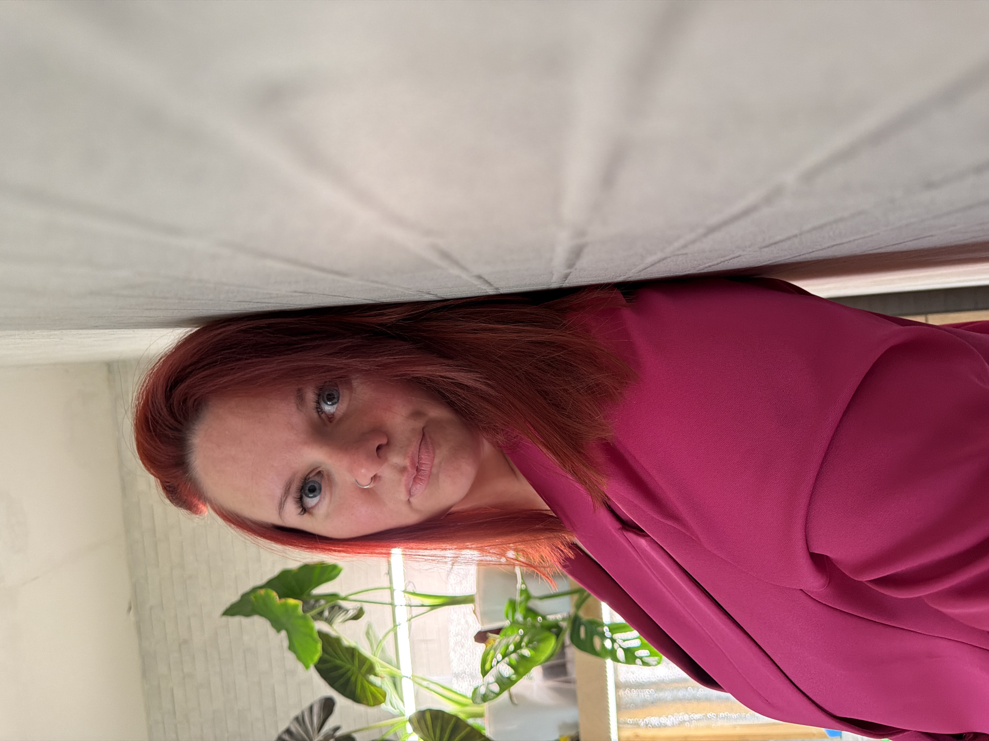 A woman with red hair and blue eyes is lying down with her head near a white wall. She is wearing a pink or magenta colored shirt or blouse. In the background, there are plants, one with large leaves, in front of a window. The location is not identifiable as the city of Chaikovsky, Russia.