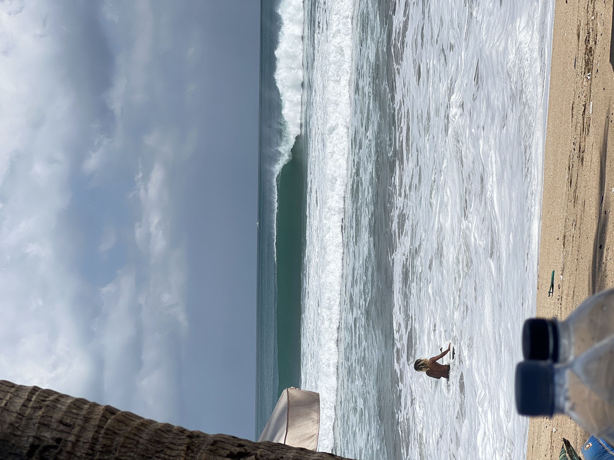In Kuta, Indonesia, on a cloudy day, a person is in the ocean with a surfboard. The person is facing away from the camera, in the water up to their waist, with the surfboard in front of them. In the background, large waves are breaking, with one wave forming a barreling shape. The water is a deep teal color. In the foreground, two plastic water bottles with blue and black caps are visible on a sandy beach. A part of a palm tree trunk is on the left side of the image, and a light-colored umbrella is behind it, partially obscuring the view of the ocean. The sky is overcast with dark clouds.