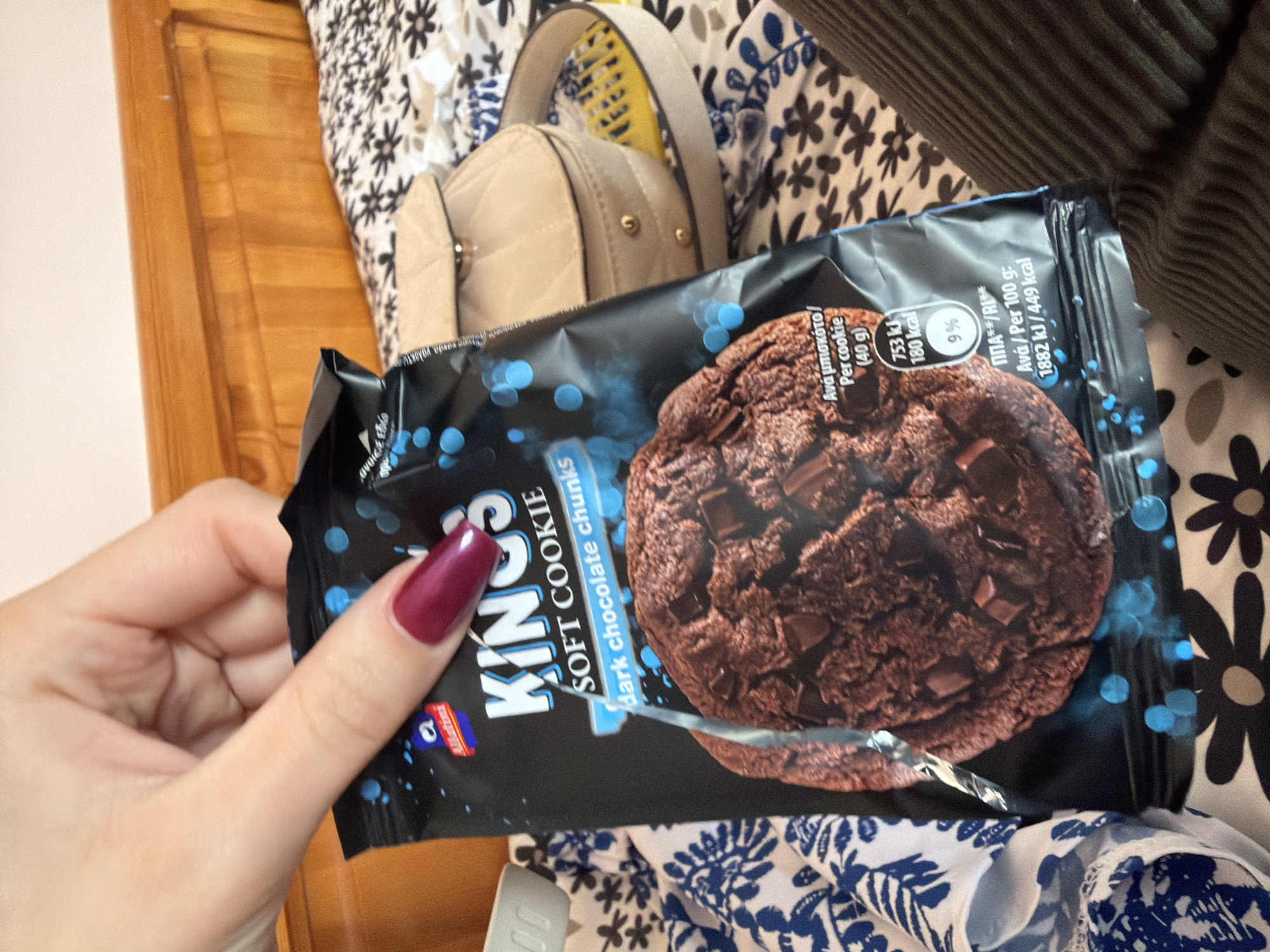A person's hand, adorned with long, dark red polished nails, holds a black package of "KINGS SOFT COOKIE dark chocolate chunks." The package features a large, appetizing image of a dark chocolate cookie with numerous chocolate chunks.
Visible text on the packaging includes the product name and nutritional information: "KINGS SOFT COOKIE dark chocolate chunks," "Ανά μπισκότο/ Per cookie (40g) 753 kJ 180 kcal 9% ΠΠΑ**/RI**," and "Ανά / Per 100 g: 1882 kJ/449 kcal." The presence of both Greek and English text for the nutritional details is notable. A small "Ballini" or "Batini" logo and another blue and red logo, likely "Allatini," are also visible on the package.
The scene is set indoors. In the background, a light-colored wooden surface, resembling a bed headboard, is seen on the left. A beige quilted handbag with a gold-colored strap and accents is positioned behind the package. Various patterned fabrics surround the objects, including a black and white floral print and a dark, vertically striped fabric, suggesting bedding or clothing. The image captures a casual moment, possibly in a bedroom, with the person about to enjoy the cookie. The location context indicates this photo is from Varna, Bulgaria.