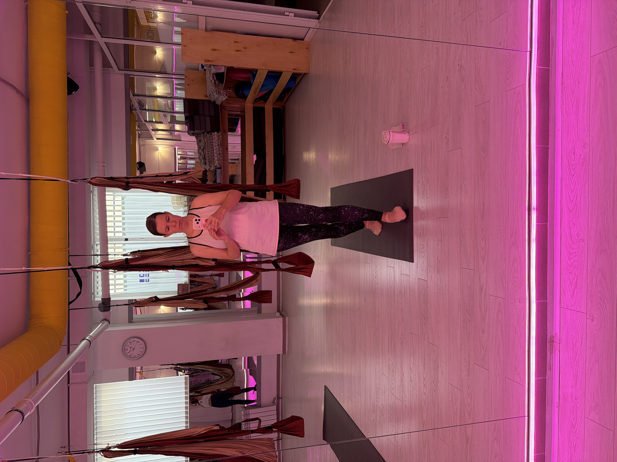 A woman is taking a selfie in a yoga studio. She is wearing a white tank top and patterned leggings, and is holding a pink phone. In the reflection of a large mirror, we see the studio filled with aerial yoga hammocks. The room is lit with pink and yellow lights. A clock is visible on the wall, showing approximately 12:09. There are yoga mats on the floor, and shelves stocked with yoga props. The floor is made of light-colored wood. The location is not specified as being in the city of Chaikovsky, Russia.