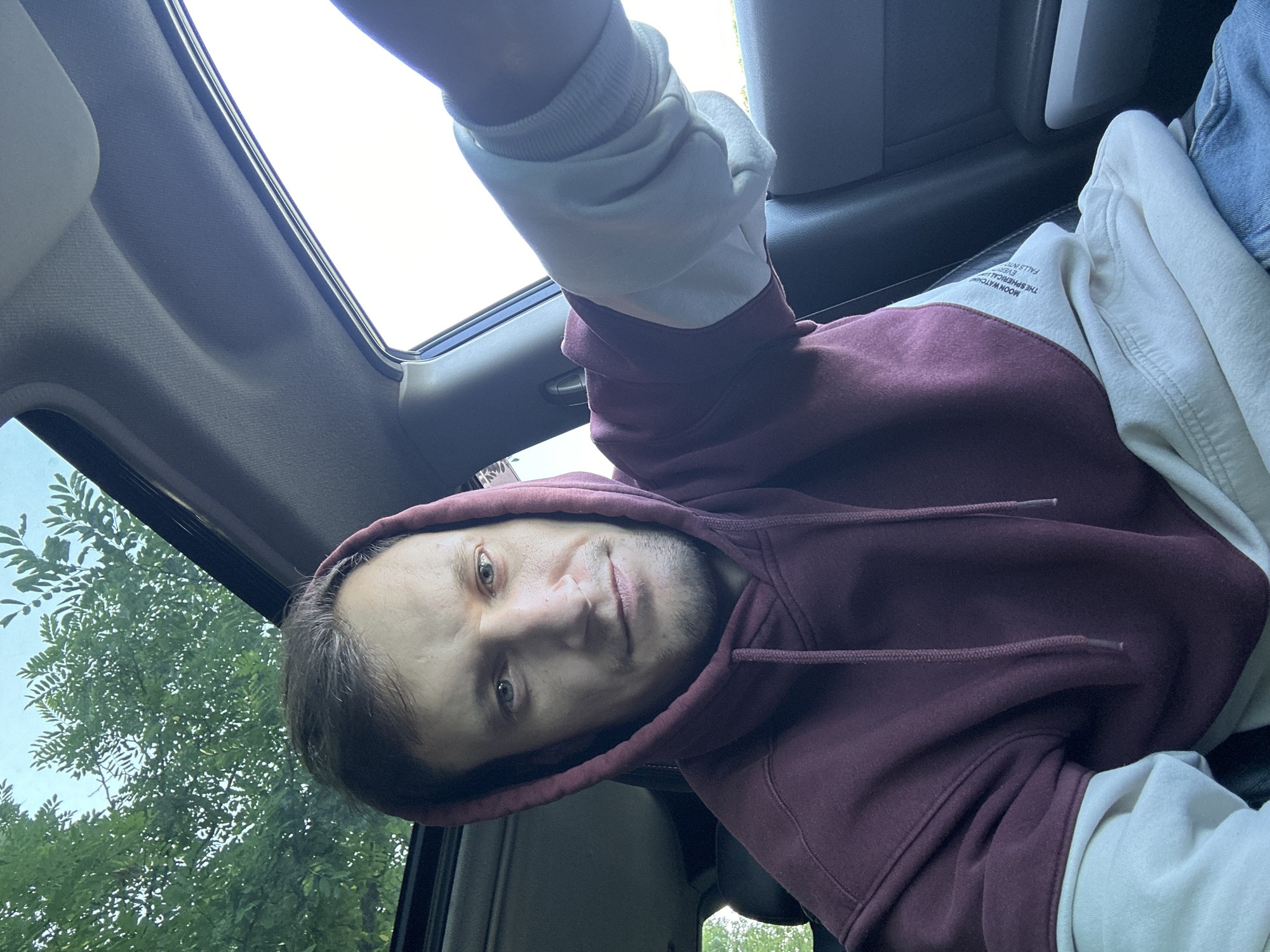 A man, appearing to be in his 30s, is taking a selfie inside a car. He is wearing a maroon hoodie with off-white sleeves and is seated in the driver's seat. The car has a sunroof open, revealing green foliage and a bright sky above. The man has short brown hair, light blue eyes, and a light beard. He is looking directly at the camera with a neutral expression. The interior of the car is visible, including the dashboard and part of the passenger seat. The exterior scene outside the car is blurred, suggesting the vehicle is in motion or parked. The man is located in Makiv, Ukraine.