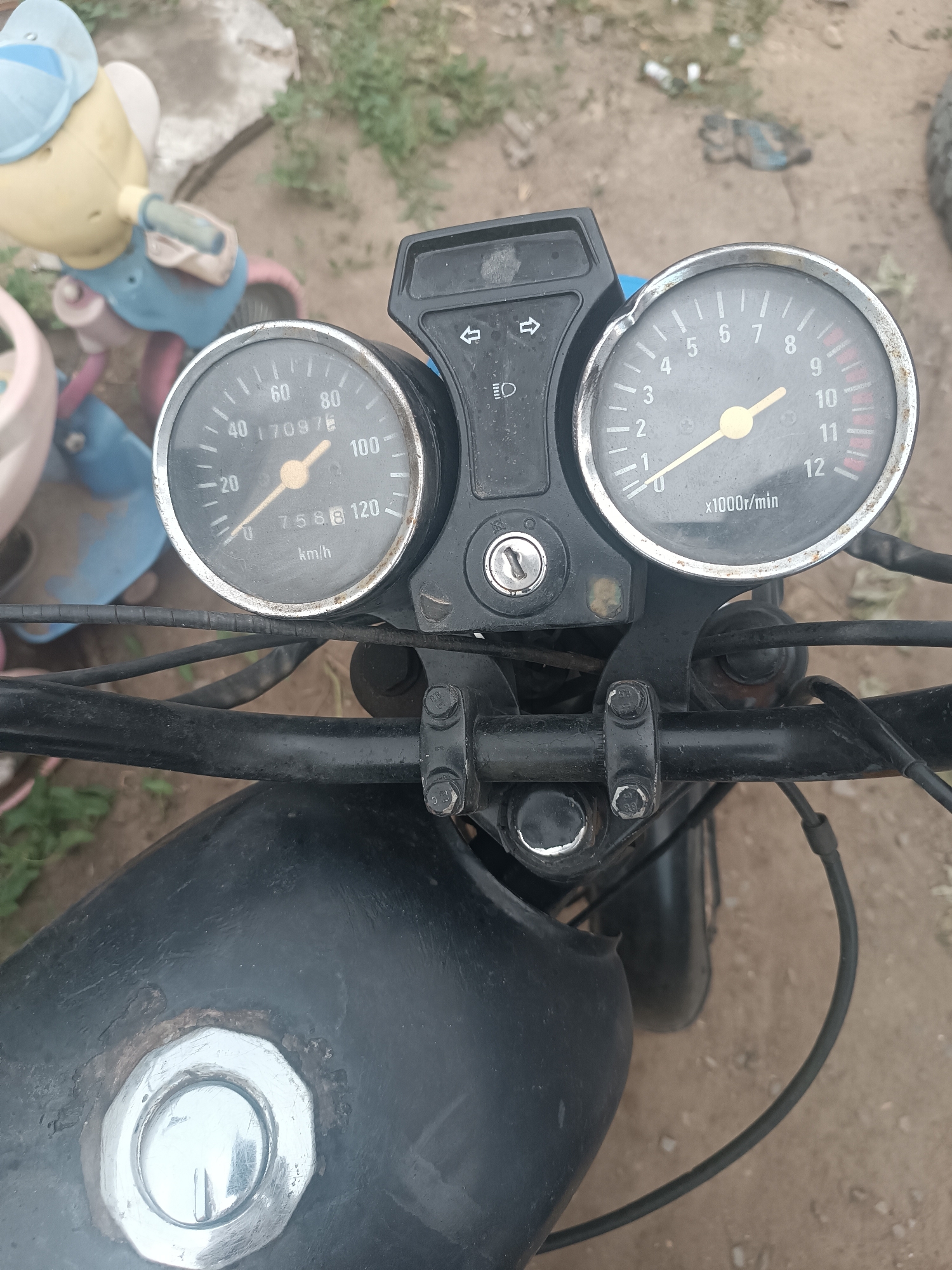 A close-up, top-down view of a motorcycle's handlebars and instrument cluster is captured. The handlebars are black and appear somewhat weathered. Attached to the handlebars is a black plastic housing containing two circular gauges. The left gauge is a speedometer, marked with increments from 0 to 100 km/h, with a needle pointing between 60 and 80. The odometer reads 70976. The right gauge is a tachometer, marked with increments from 0 to 12 (representing x1000r/min), with a needle pointing between 0 and 1. To the left of the gauges is a silver ignition keyhole. Above the gauges are indicator light symbols for headlights and turn signals. The motorcycle's black fuel tank is partially visible in the lower portion of the frame, featuring a silver cap. In the blurred background, a child's blue and yellow toy, possibly a tricycle, and some greenery are discernible. The ground appears to be dirt or sand. The scene is outdoors, and the lighting suggests daylight. There are no people present in the image. The image is taken in Klyuchi, Russia.