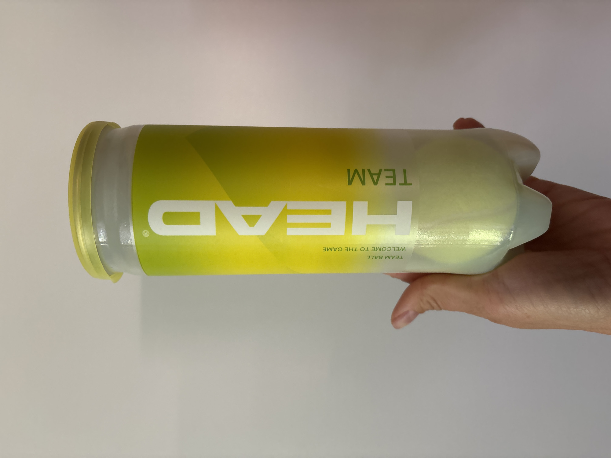 A human hand with light skin tone is holding a clear plastic cylindrical container of three tennis balls, positioned vertically. The container features a light yellow translucent plastic lid and a molded bottom designed to cradle the balls. A prominent label on the container displays the brand "HEAD" in large white vertical lettering against a lime green and yellow gradient background. Subordinate text on the label includes "TEAM BALL," "WELCOME TO THE GAME," and "TEAM." Three fluorescent yellow-green tennis balls are visible inside the container. The background consists of a plain, light-colored wall, providing a neutral backdrop. The scene is located in the city of Chaikovsky, Russia.