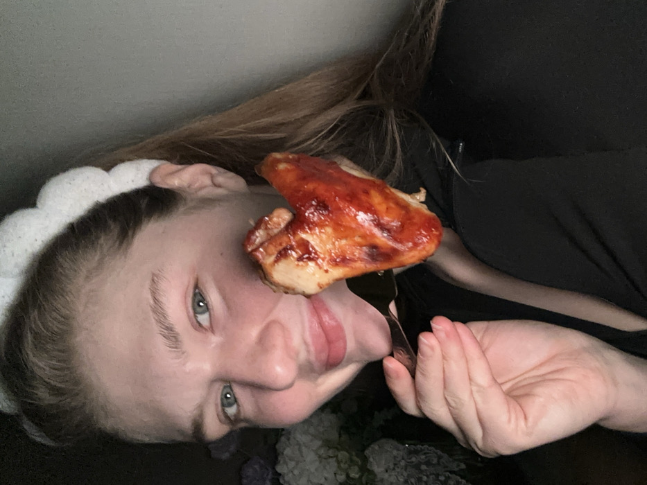 A woman, turned sideways, smiles at the camera, holding a piece of chicken coated in red sauce on a fork near her face. She has light skin, blue eyes, and light brown hair with a white, cloud-like headband. She wears a black shirt. A floral arrangement with white flowers sits near her hand.