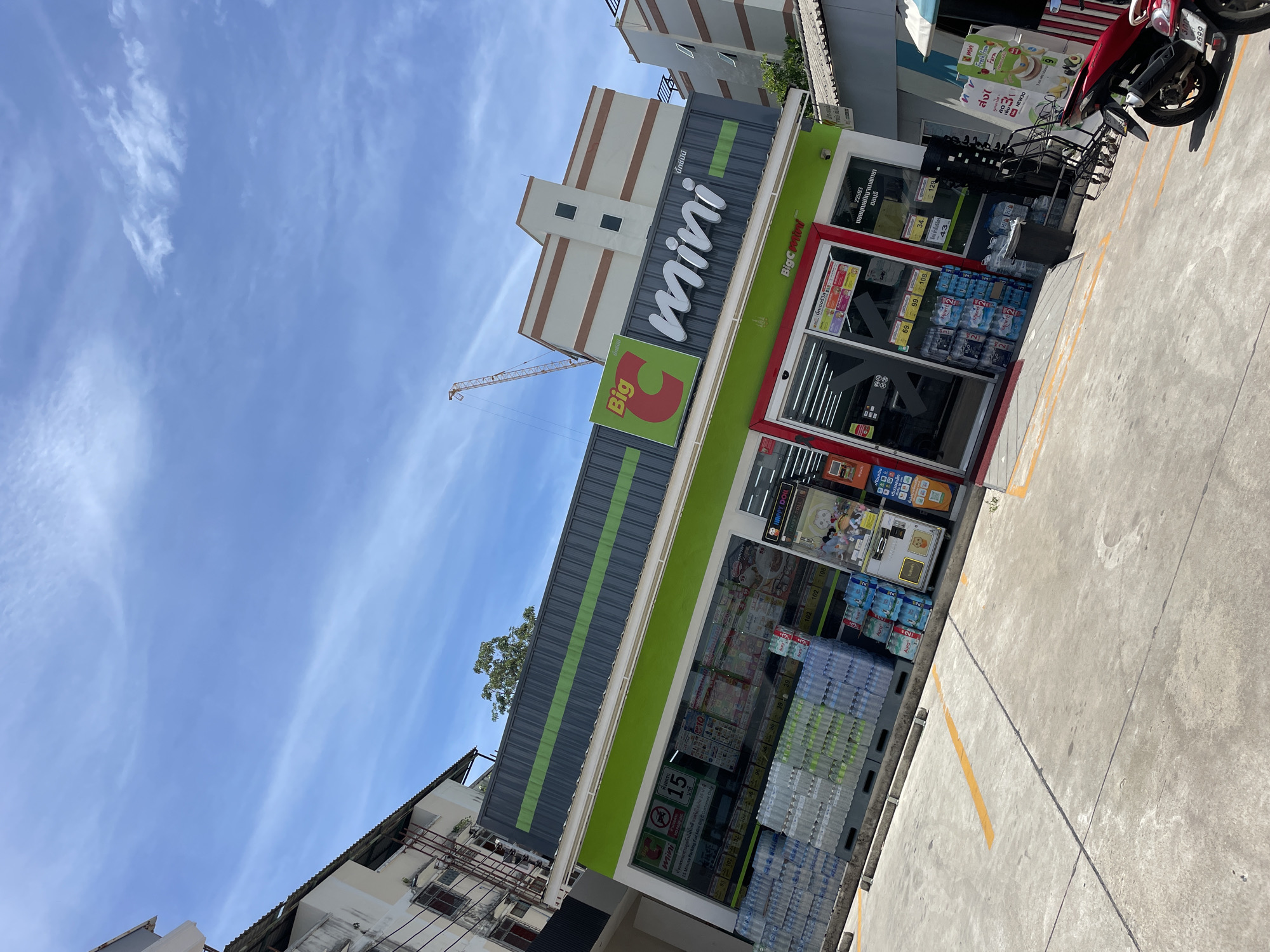 This is a medium-angle, eye-level, full shot of a Big C Mini convenience store in Pattaya City, Thailand, on a bright, sunny day. The sky is blue with scattered white clouds and a yellow construction crane is visible in the background. The store is a modern, single-story building with a green and gray facade. The name "Big C mini" is prominently displayed in white lettering on the front of the store. The storefront features large glass windows showcasing shelves stocked with bottled water and other merchandise. Outside the store, there are stacks of bottled water, a trash can, shopping carts, and a red motorcycle parked to the right. Yellow painted lines on the concrete ground indicate parking spaces. Adjacent buildings, including a multi-story apartment building, are visible on either side of the convenience store. The overall scene depicts a typical urban retail environment in Pattaya City.
