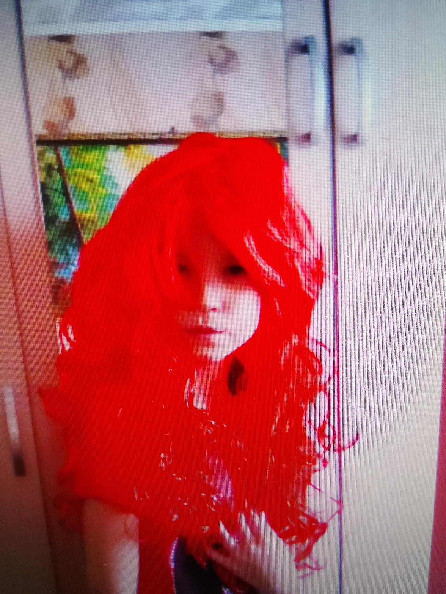 A low-angle, slightly blurry, full-shot photograph depicts a child wearing a vibrant, voluminous red wig. The child's face is partially visible, with their eyes appearing to be closed or looking down. The wig obscures most of their features and extends outwards, framing their face and shoulders. Behind the child, a closet door is visible, with two handles. The closet interior contains what appears to be a picture or tapestry with a colorful, leafy pattern. The image quality is poor, likely due to being a photograph of a screen.