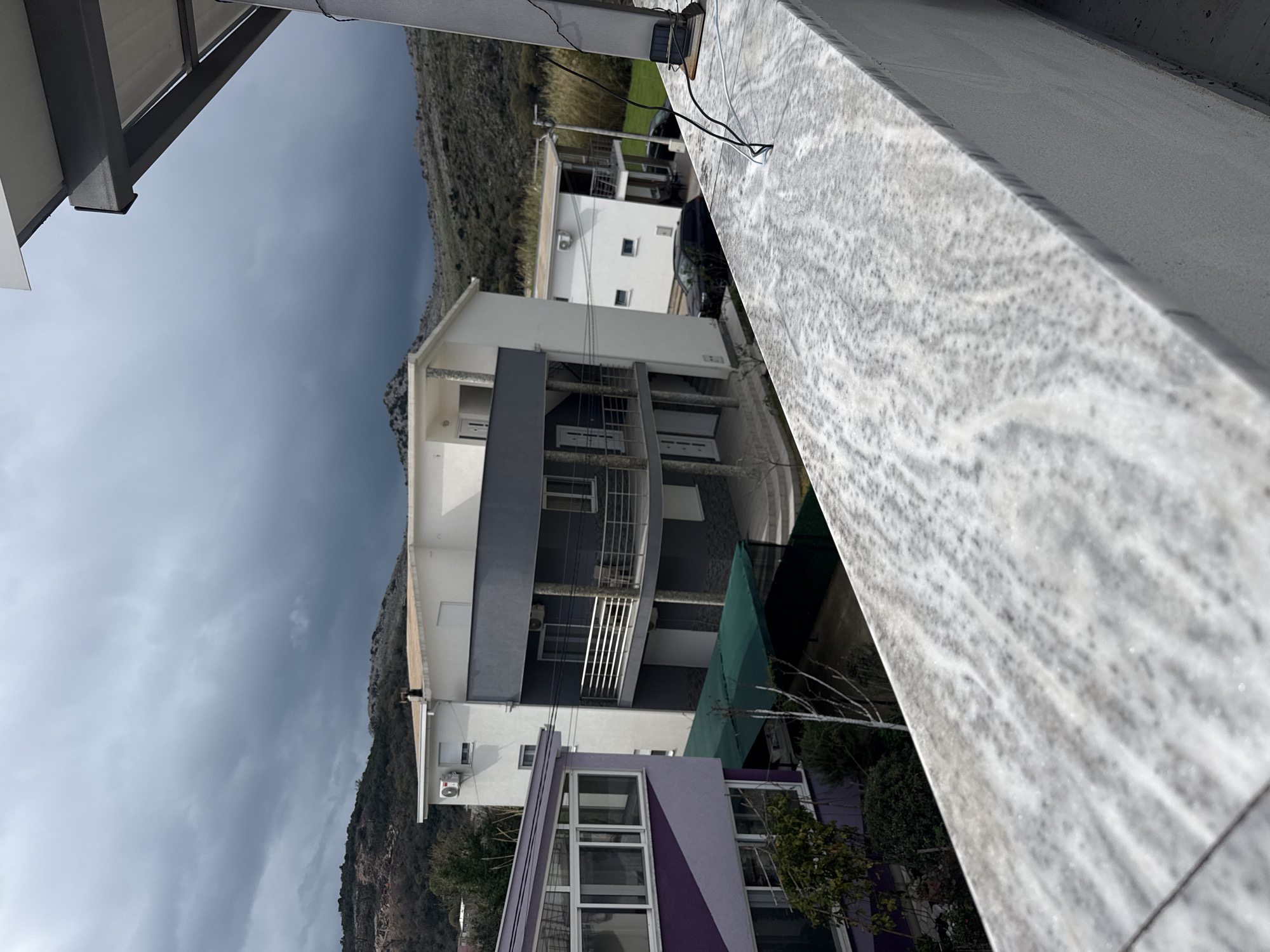The image depicts an outdoor scene observed from an elevated balcony, featuring residential buildings against a mountainous backdrop under a heavily overcast sky. In the immediate foreground, a light-colored, possibly marble or granite, balcony railing runs horizontally across the bottom of the frame, with gray and white veining. On the right edge of this railing, several black and white electrical wires are visible, connected to a small, dark rectangular object.
The midground features multiple multi-story residential structures. To the left, a building with a distinct purple and white facade includes an upper floor with large windows, possibly an enclosed balcony. Below this, a dark green tarp provides overhead cover. In the center, a three-story building features a white upper section and a dark gray middle section with a prominent balcony adorned with a white metal railing. An outdoor air conditioning unit is mounted on the top floor of this building. To its right, partially obscured, is another white building with a balcony. A black SUV-type vehicle is parked near this rightmost building.
The background consists of rugged, vegetated hills and mountains with exposed rock formations. The sky above is entirely covered by dark gray, dense clouds, indicating an overcast or stormy weather condition. No people are visible in the scene. The overall environment appears to be a residential area at the base of mountains, likely in the city of Unknown, Unknown.