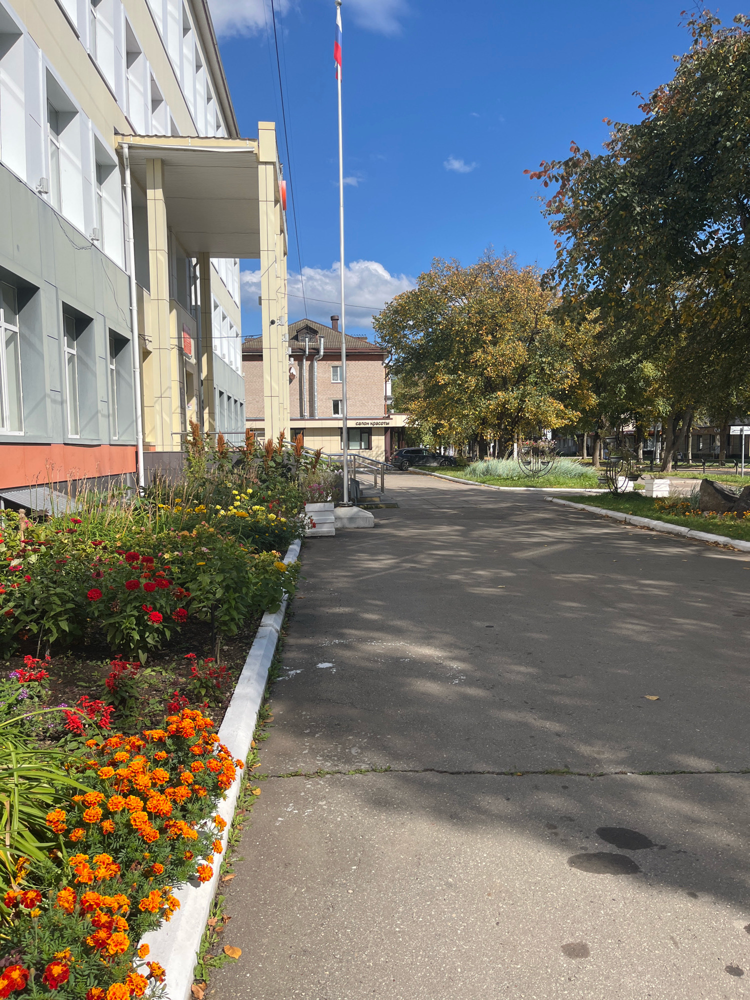 The image depicts the entrance area of a building in Chaikovsky, Russia. A light-colored, multi-story building with many windows occupies the left side of the frame, featuring a covered entrance supported by columns. A vibrant flower bed with red and orange flowers runs along the building's base, bordering a paved walkway. Beyond the building and flower bed, a paved road stretches into the distance, lined by trees with autumn foliage. A tall flagpole with the Russian flag is visible near the building. In the background, other buildings and more trees contribute to a suburban or town-like setting. The sky is blue with scattered clouds, indicating fair weather. There are no people visible in the image.