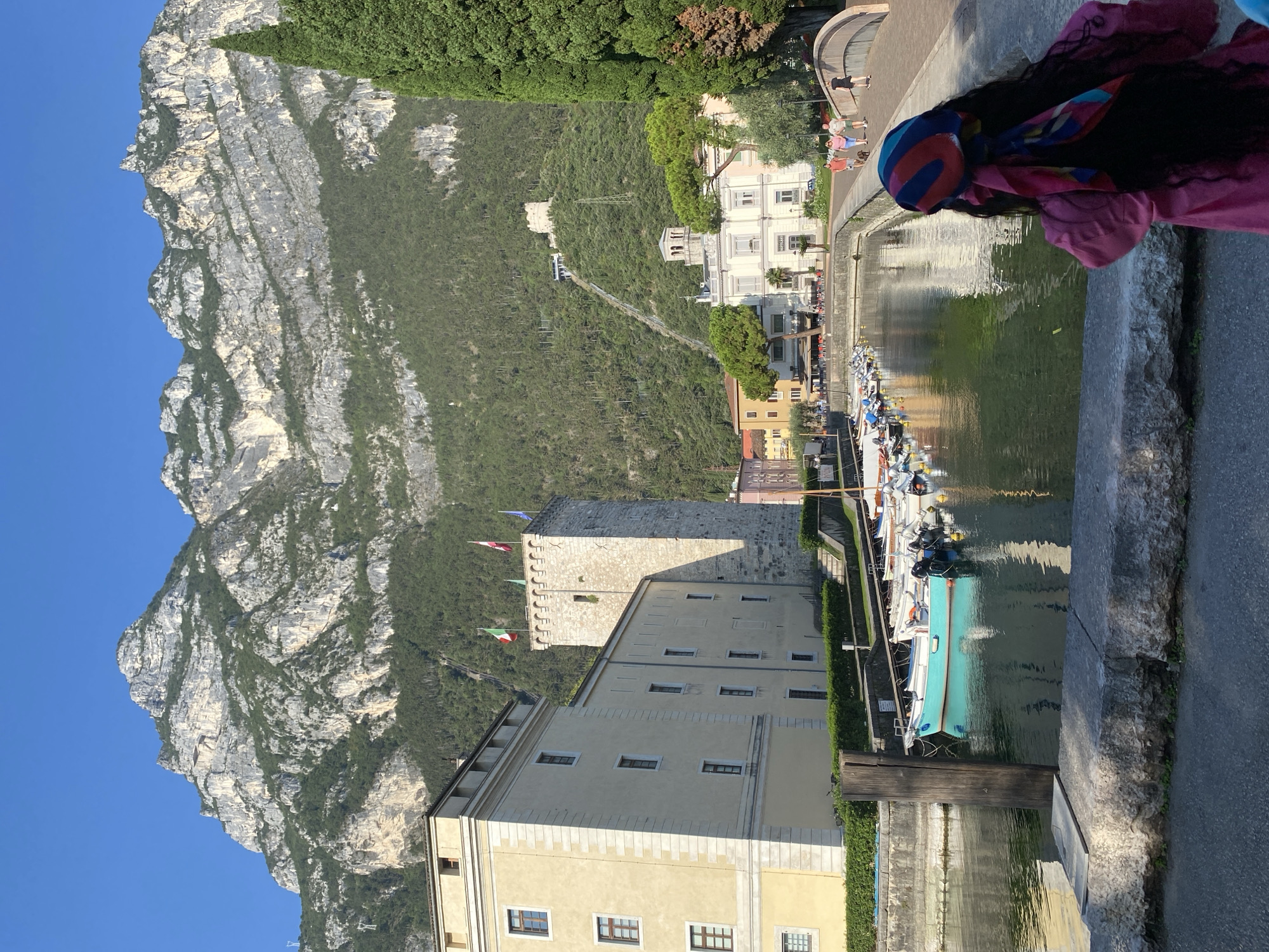 The image captures a view of the waterfront in Riva del Garda, Italy, looking towards the mountains. In the foreground, a person with long dark hair wearing a pink outfit and a colorful headscarf faces away from the camera, looking towards the water. A stone wall separates the person from a paved walkway.

To the left, a light-yellow building with numerous windows stands beside a tall, stone tower topped with flags. Boats are docked in a narrow canal in front of these structures, with some of the boats being turquoise and white. The canal leads towards a bridge in the middle ground, crossed by several individuals.

Beyond the canal and bridge, a cluster of buildings, including a white building with arches and a smaller building with a clock tower, forms part of the town. In the background, a steep, tree-covered mountain rises dramatically under a clear blue sky. A funicular track and a small, ruined structure are visible ascending the mountainside. Cypress trees are prominent on the right side of the image. The scene is illuminated by bright sunlight, suggesting it is a clear day.