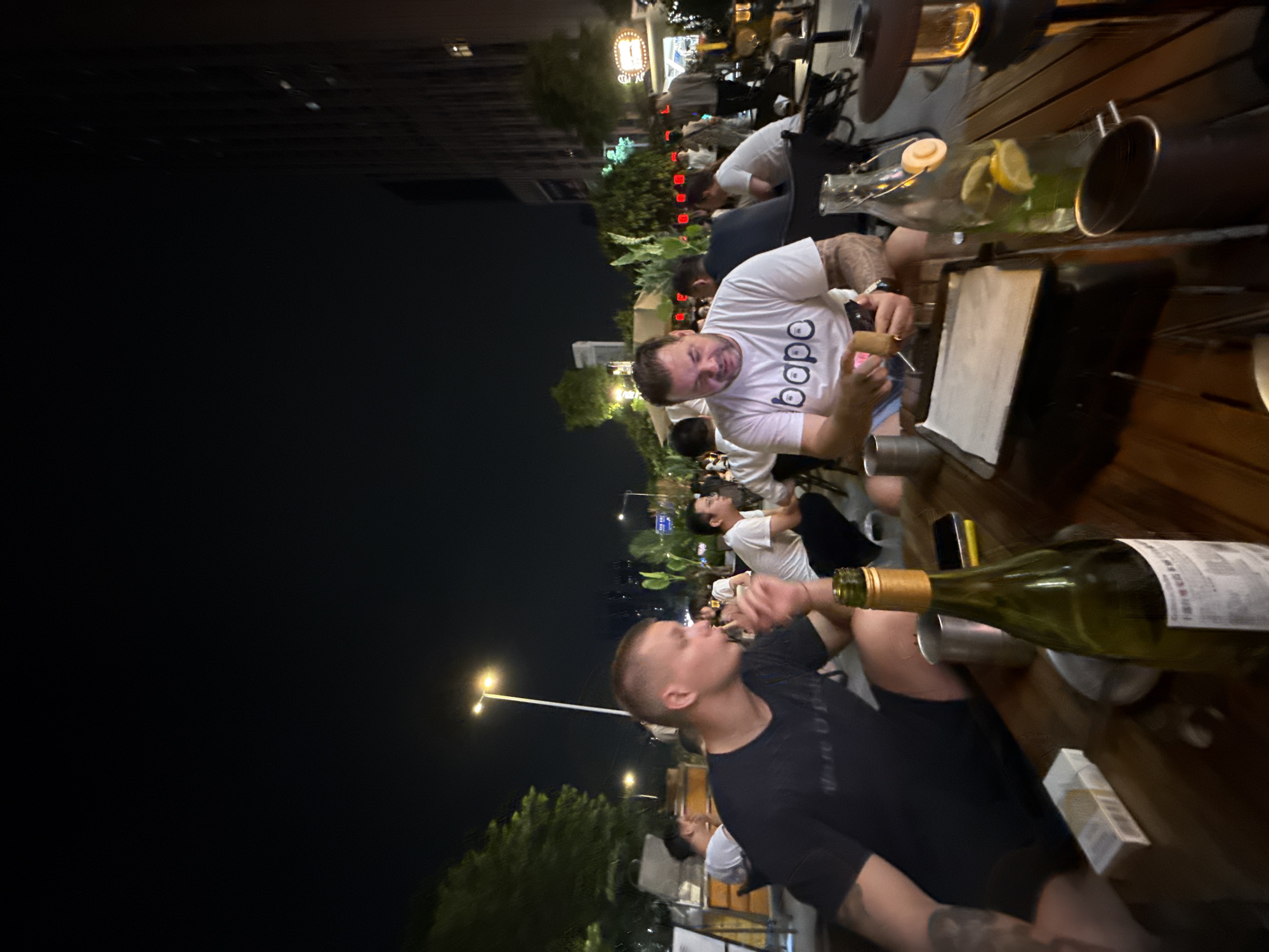 An outdoor nighttime scene depicts several individuals seated at wooden tables, appearing to be at a restaurant or bar in Haizhu District, China. In the foreground, a man in a black t-shirt is captured in profile, with a cigarette in his mouth and his hand raised towards it. Next to him, a green wine bottle stands upright on the table, alongside a pack of cigarettes and a metal cup.
To the right of the frame, another man wearing a white t-shirt with the word "bape" printed on it is actively engaged with his mobile phone and appears to be holding a small object, possibly a lighter or a tool, in his other hand. A glass bottle filled with water and lemon slices is positioned on the table in front of him, along with a metal cup. Behind these two men, other patrons are visible, some seated and some standing, in a dimly lit environment with plants and city buildings in the background. The overall atmosphere suggests a social gathering.