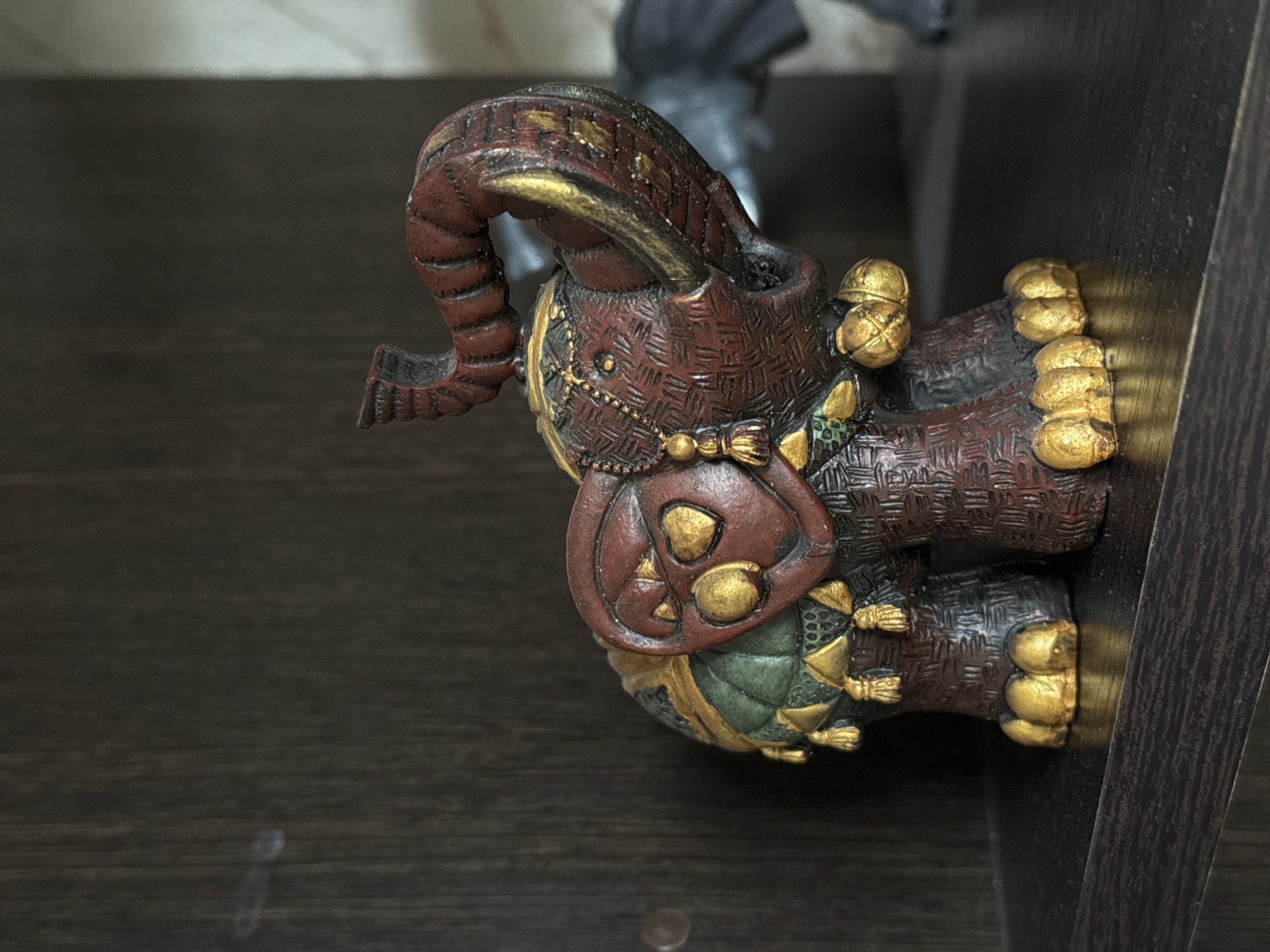 A close-up shows a decorative, reddish-brown elephant figurine adorned with gold accents, situated on a dark brown wooden surface. The elephant is equipped with a saddle and elaborate headgear. The texture of the figurine suggests it might be made of resin or a similar material with intricate detailing. The setting seems to be indoors, possibly on a shelf or piece of furniture. The city of Novo-Nikol'skoe, Russia, is not discernible from the image.
