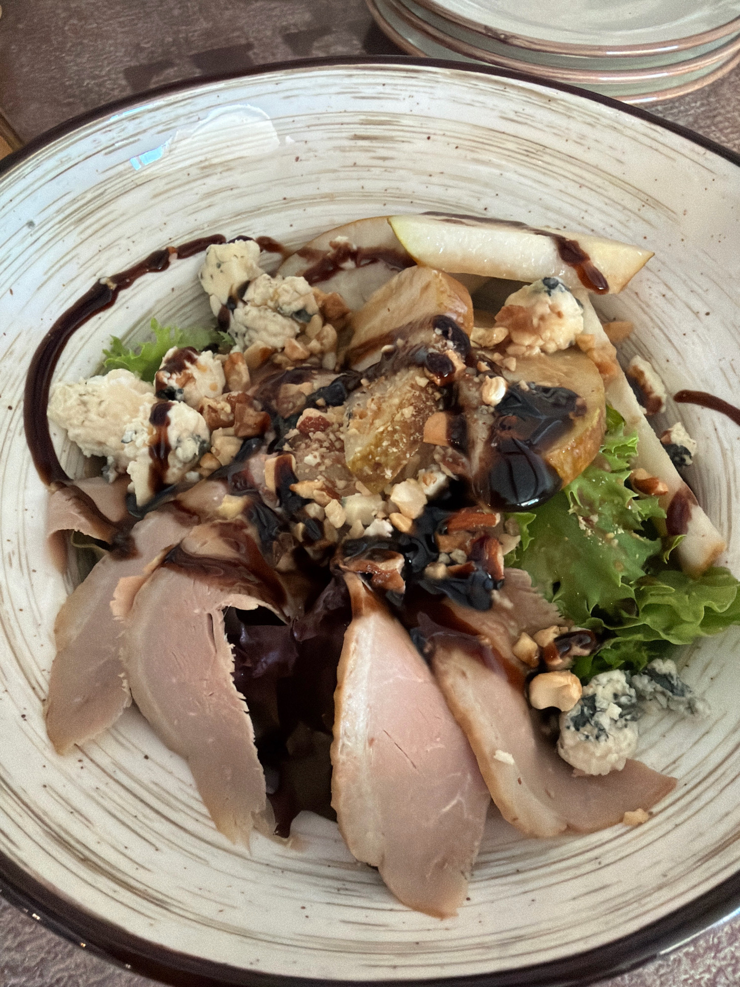 A close-up, overhead shot shows a salad in a rustic, ceramic bowl with brown and white concentric markings. The salad is composed of thinly sliced roast meat, pear slices, crumbled blue cheese, chopped nuts, and leafy green lettuce. A dark, viscous dressing, likely balsamic glaze, is drizzled over the ingredients. The bowl is placed on a textured, brown surface, possibly a wooden table. In the background, a stack of similar ceramic plates is visible. The lighting is natural and appears to be from an overhead source. The image is sharp and well-lit, highlighting the textures and colors of the food. The scene depicts a prepared meal, likely in a restaurant setting.