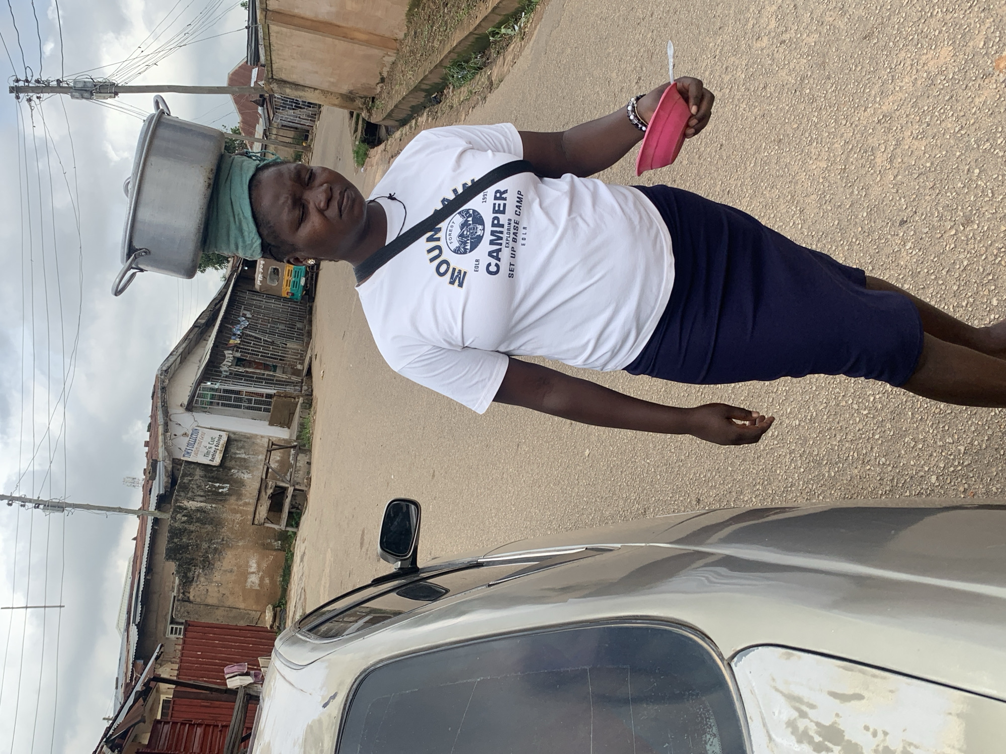 In Jos, Nigeria, a woman walks beside a silver car. She has a large metal pot balanced on her head, cushioned by a green cloth. The woman is wearing a white t-shirt with a "Mountain Camper" logo and a dark skirt. In her right hand, she holds a pink bowl with a spoon. The background shows a street lined with buildings, some with corrugated metal roofs. A utility pole with wires is visible, and a yellow and blue tricycle is parked further down the street. The sky is overcast.