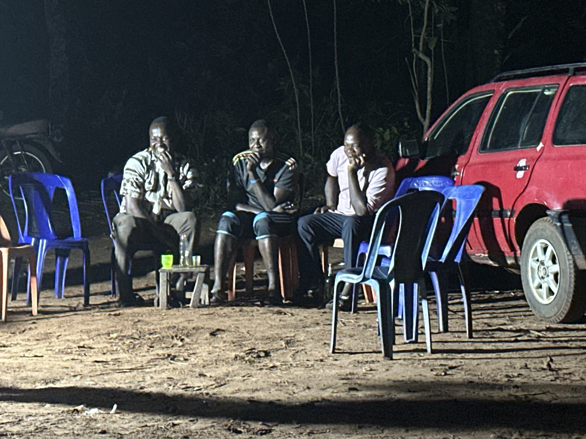 Three men are seated on chairs in an outdoor setting at night. They are arranged in a row, with two plastic blue chairs in front of the rightmost man. A small wooden table with glasses on it sits between the first two men. To the right of the group is a red SUV. Behind the men, the darkness obscures details, but there appears to be a tree and a glimpse of a motorbike. The ground is dirt. The location is the city of Unknown, Unknown.