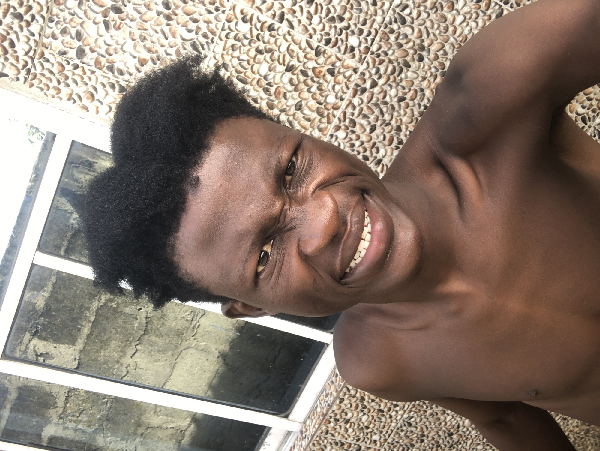 A close-up selfie captures a young Black male from the chest up, outdoors. He has dark, curly hair and is bare-chested, displaying dark brown skin. His facial expression is a scrunched-up grin, revealing his teeth. He is positioned in front of a wall covered in small, round, tan and brown stones, and to his left, a window with a brick wall visible behind it. The overall lighting suggests it is daytime, with shadows indicating the sun is out. The location is identified as Ado, Nigeria.
